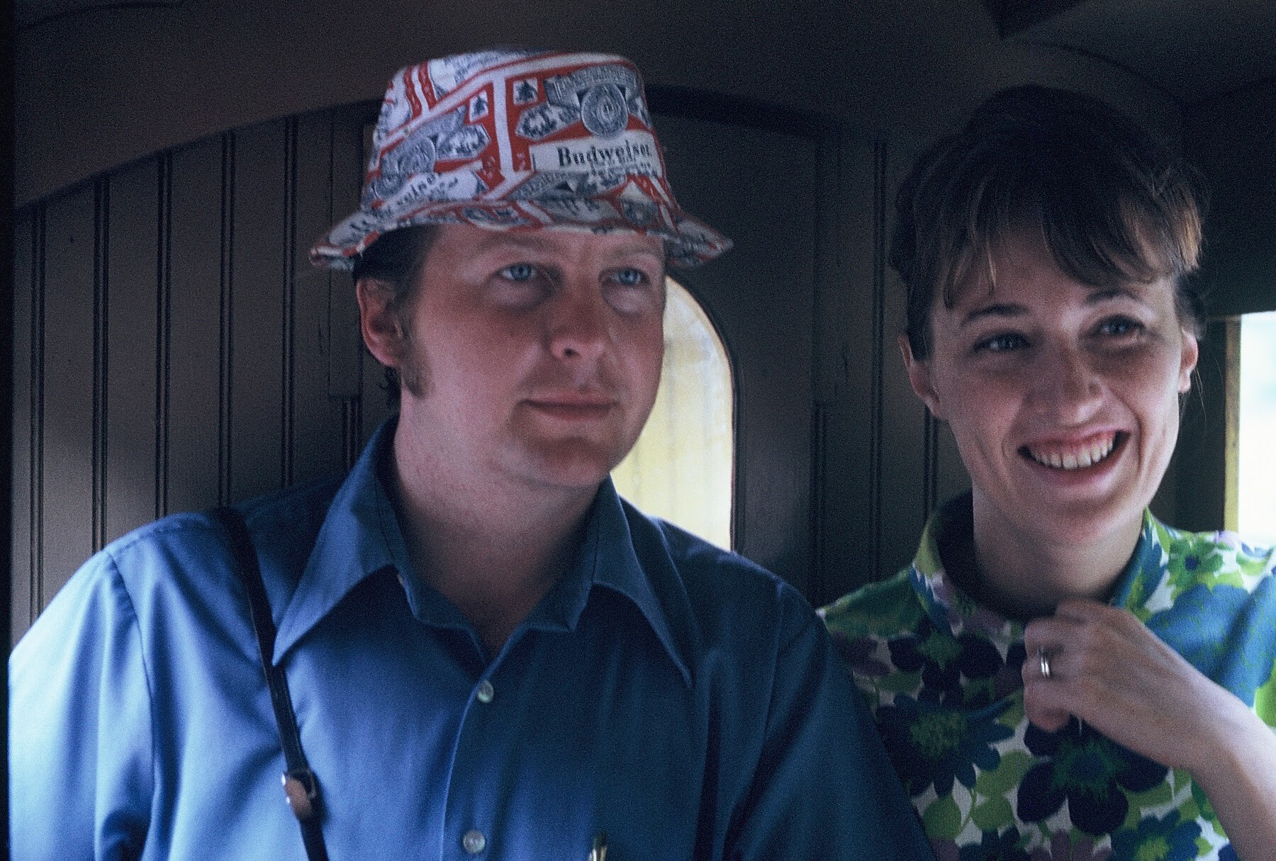 Two friends riding in a train car; man wearing a red‑white Budweiser hat looks off to the side, while a smiling woman in a green floral shirt stands next to him.
