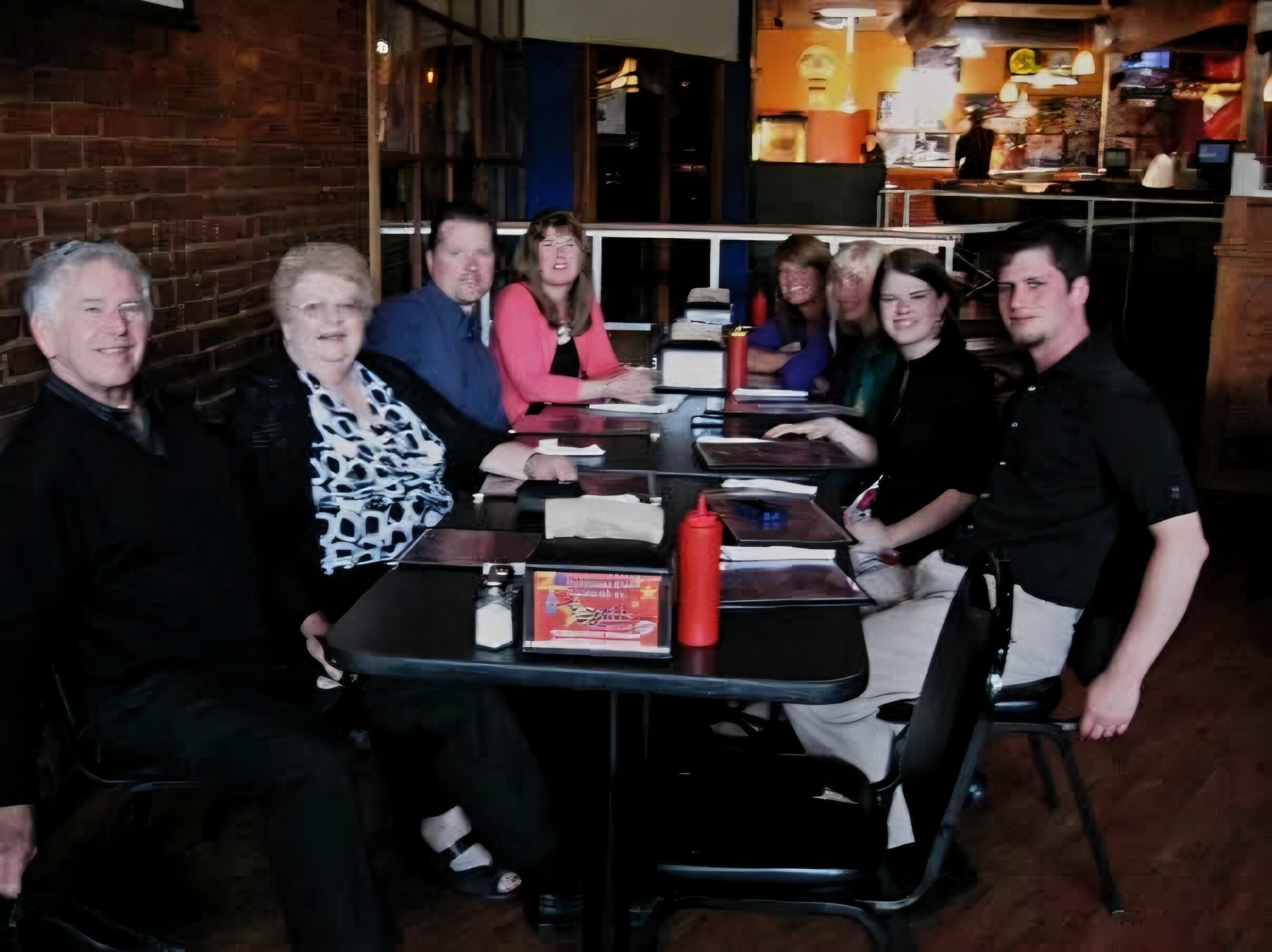 Group of adults seated around a restaurant table, some smiling, with menus and red condiment bottles on the table.