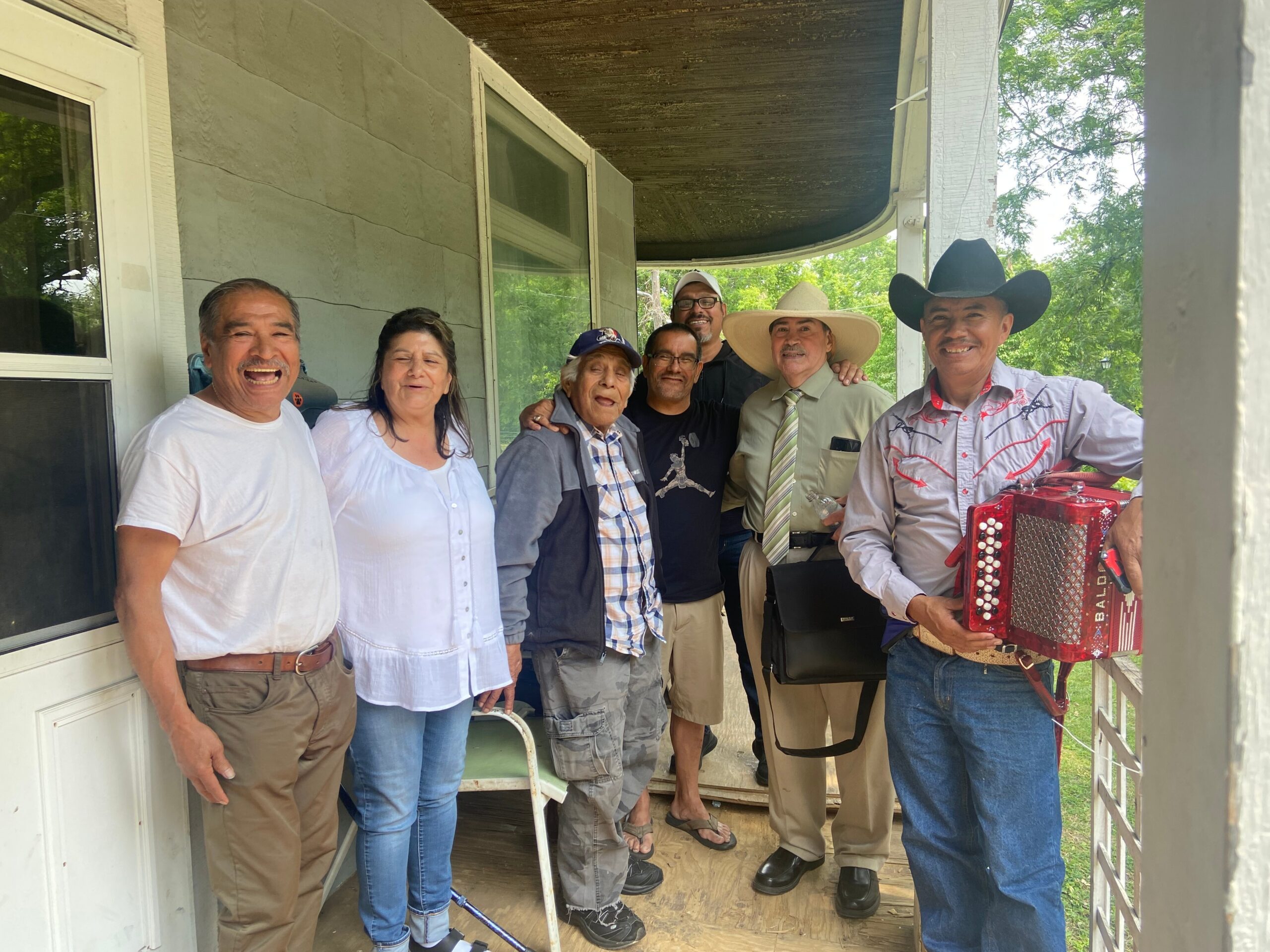 Group of adults posing on a porch, smiling; several wear cowboy hats, one person plays a red accordion. Sunny day.