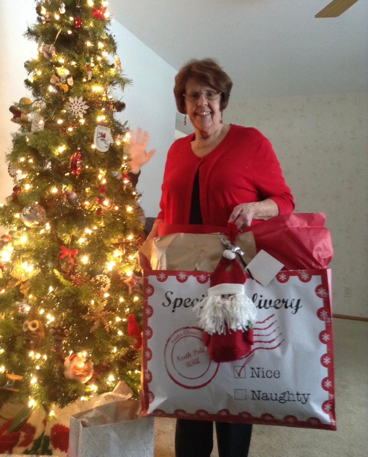Smiling woman in a red sweater waves beside a lit Christmas tree, holding a large gift bag and presents.