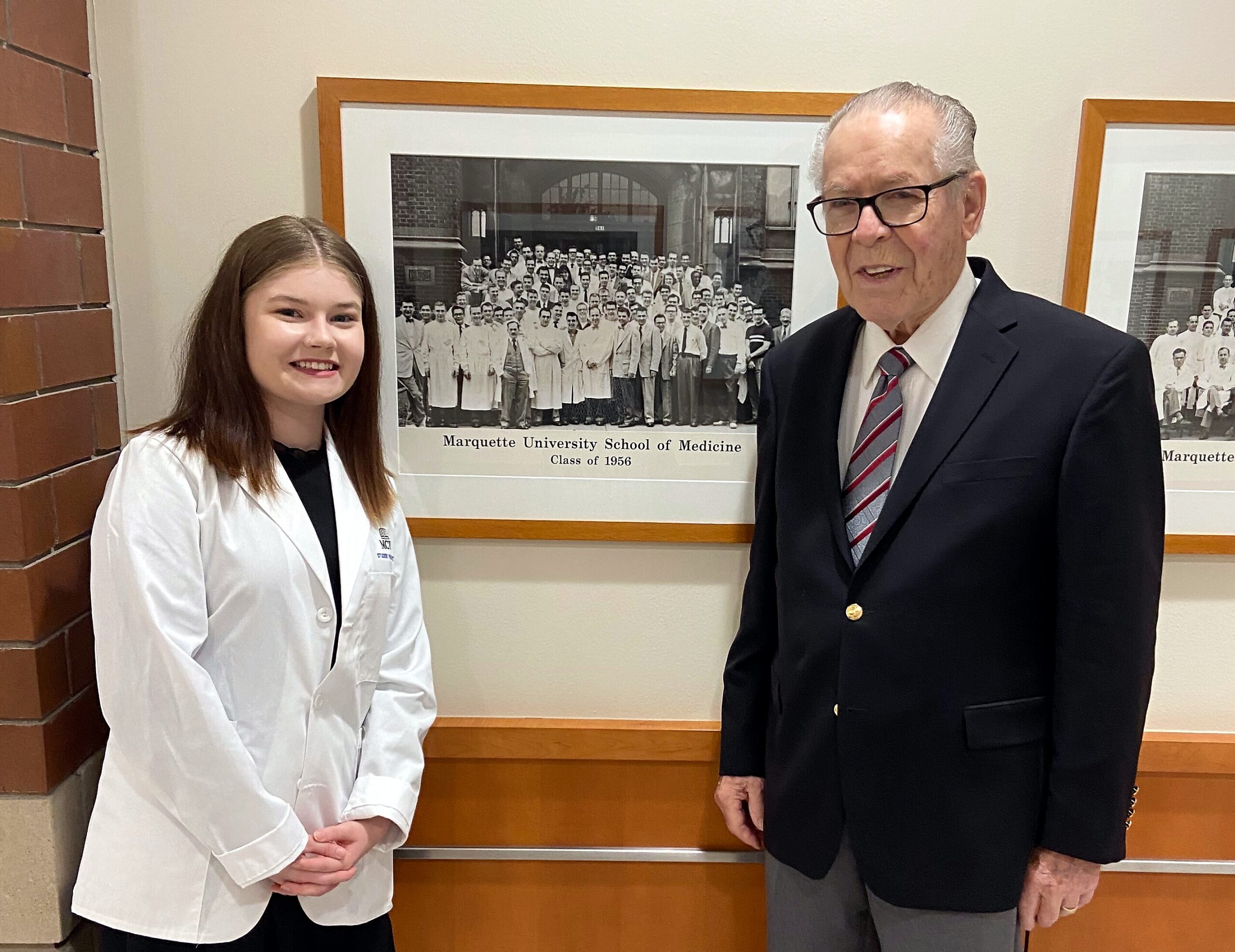 Young woman in a white lab coat and an older man in a dark suit pose beside a large black-and-white class photo framed on the wall, which reads 'Marquette University School of Medicine, Class of 1956'.