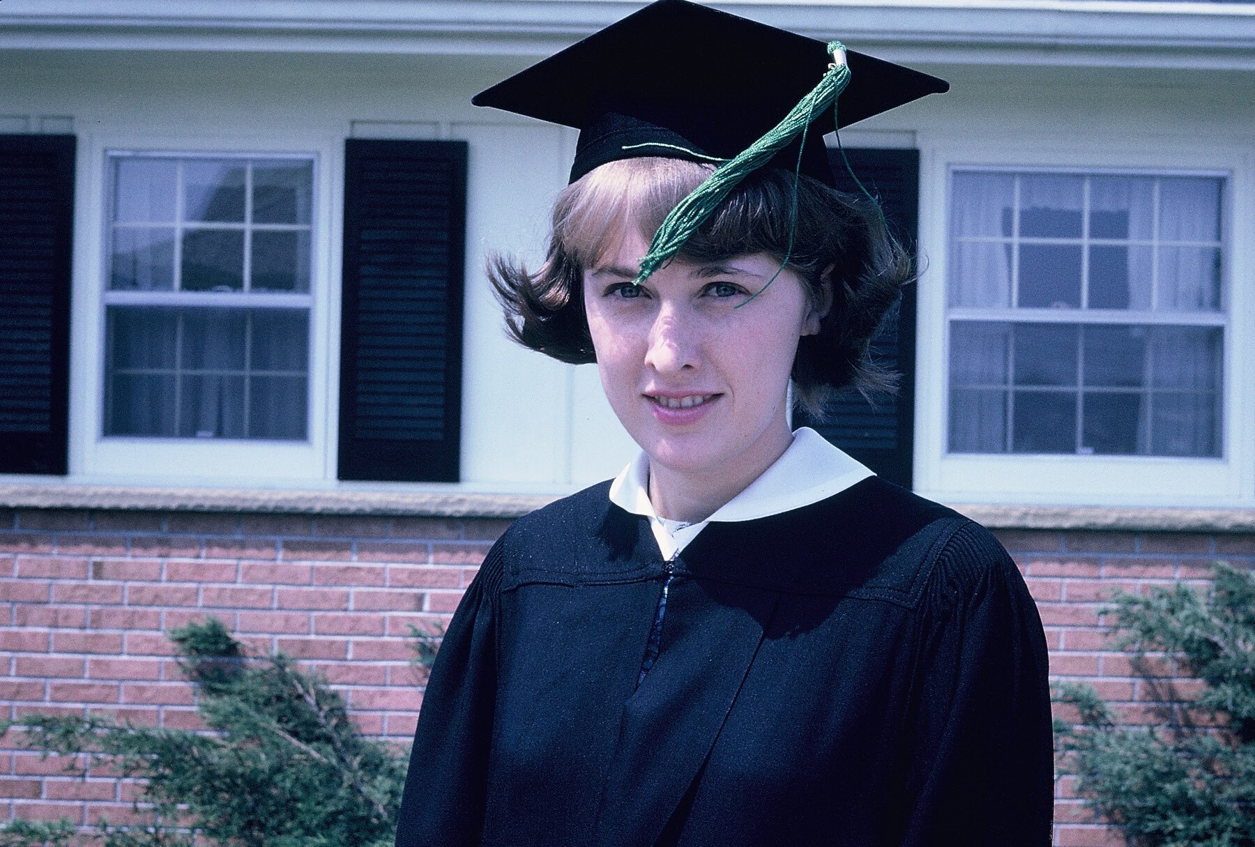 Graduate in a black cap and gown posing outside in front of a brick building with windows and shrubs.