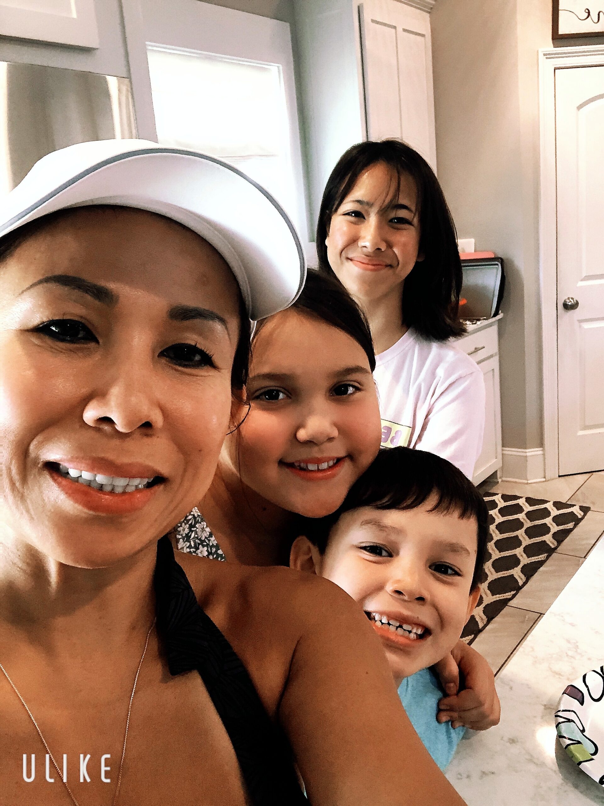 Group of smiling people taking a selfie in a bright kitchen, with a woman in a white cap in the foreground and two children behind her.