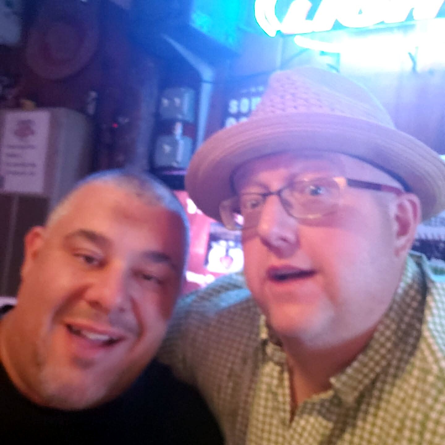 Two men posing for a selfie in a bar, one wearing a straw fedora and glasses, the other in a dark shirt.