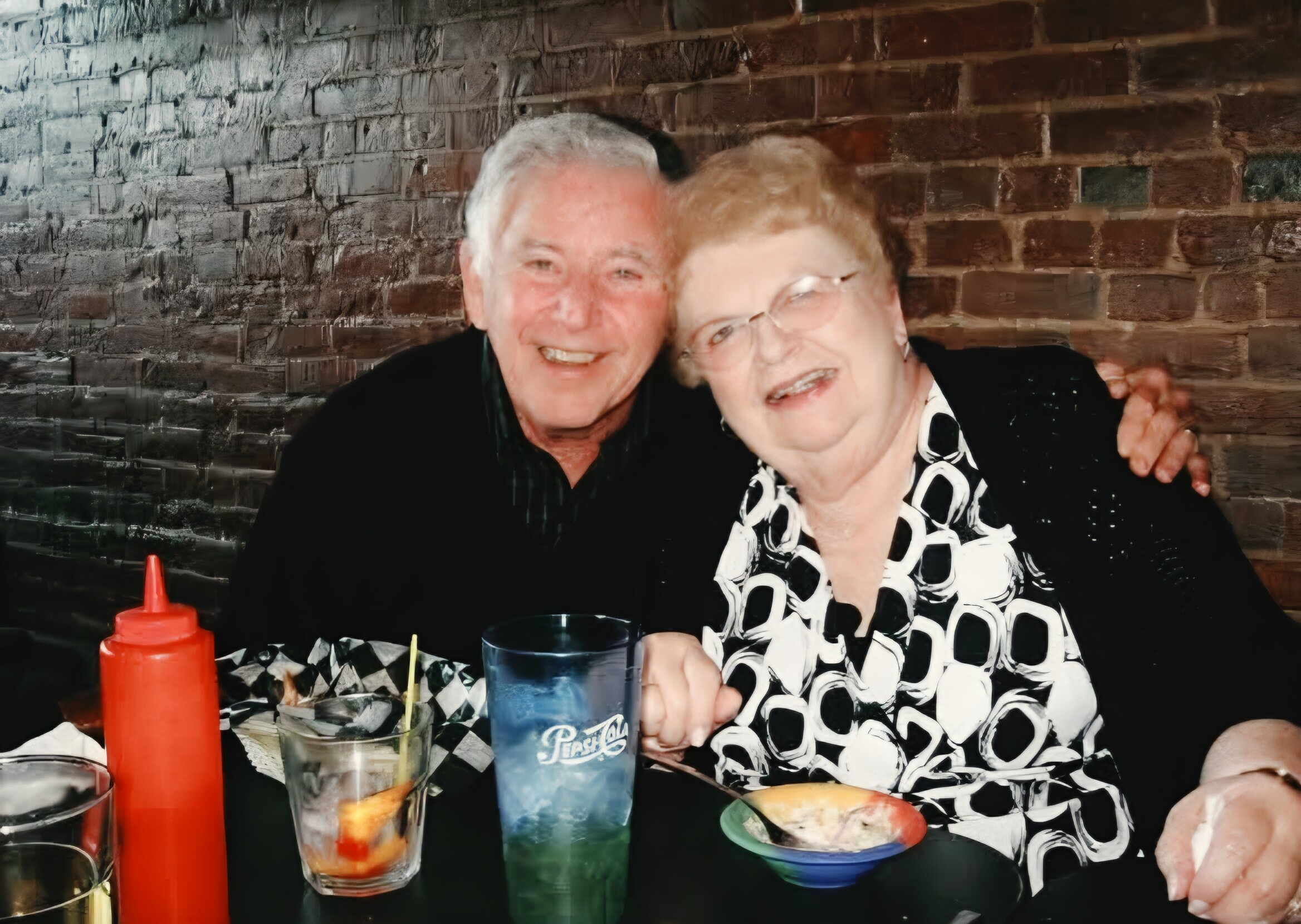 Two elderly friends smiling at a table with colorful drinks and a red ketchup bottle in front, brick wall behind them.
