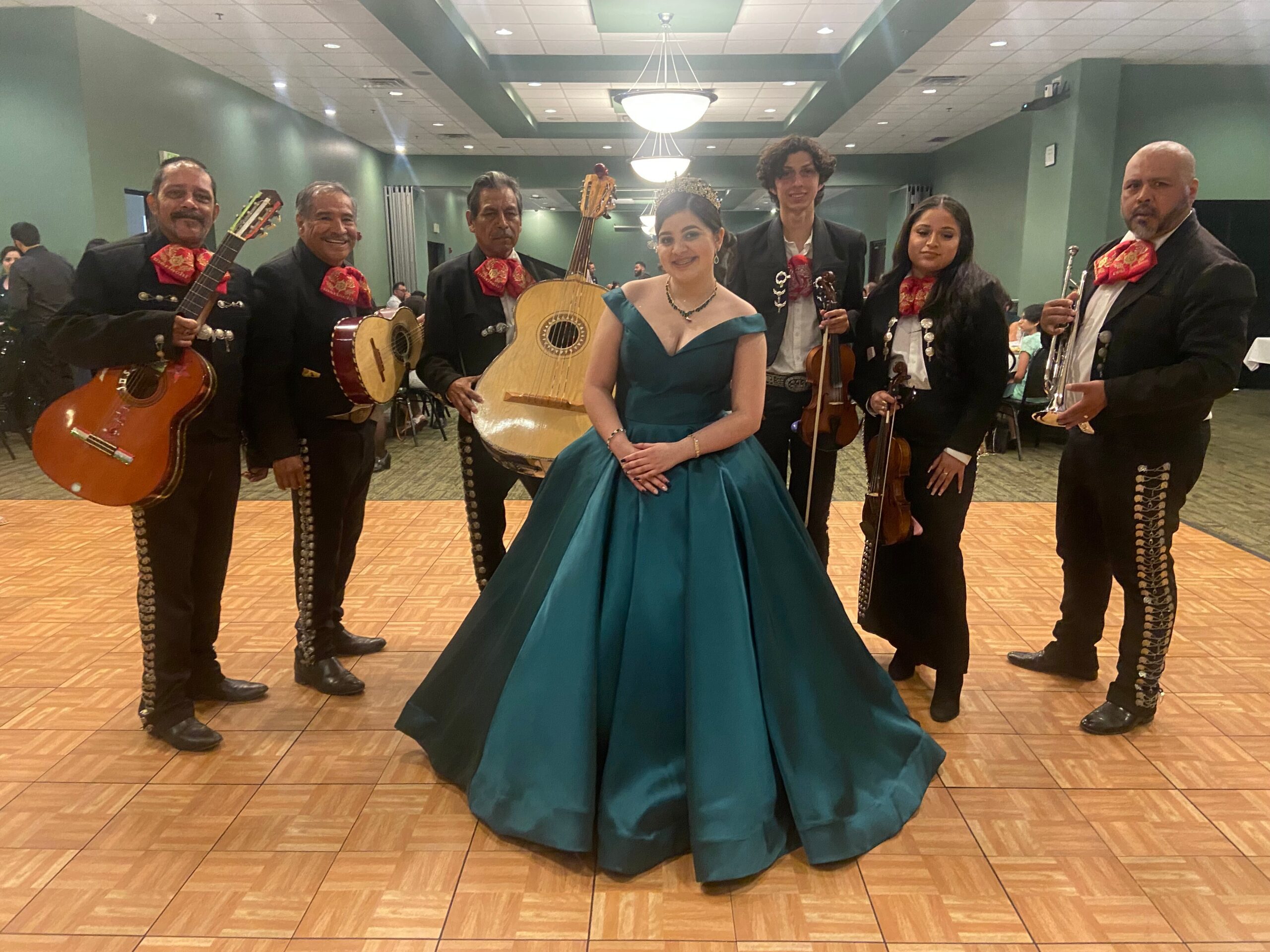 Mariachi performers in black suits with red bow ties pose with guitars and a violin around a woman in a teal evening gown in a banquet hall.