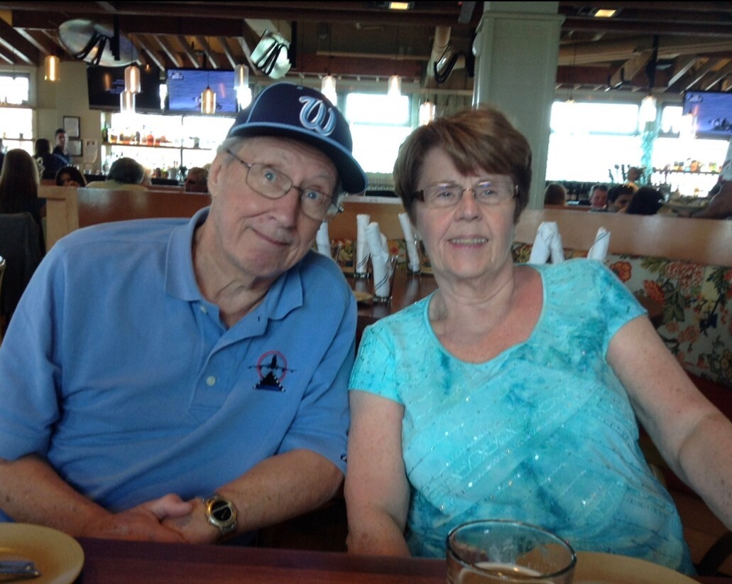 Older couple dining at a restaurant, man in blue polo and cap beside a woman in turquoise top at a booth, smiling at camera.