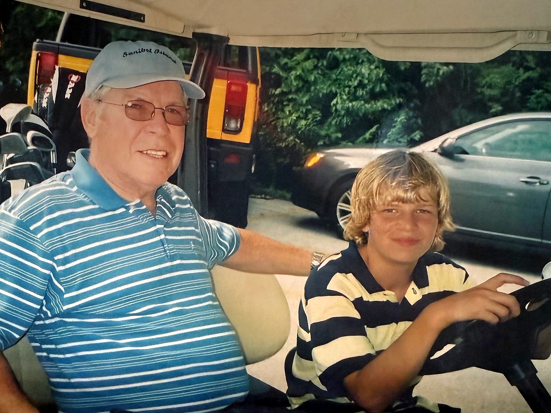 Two people riding in a vehicle, the older man wearing a striped polo and a light cap with glasses, smiling at the camera while a younger person sits beside him in a black-and-tan striped shirt.