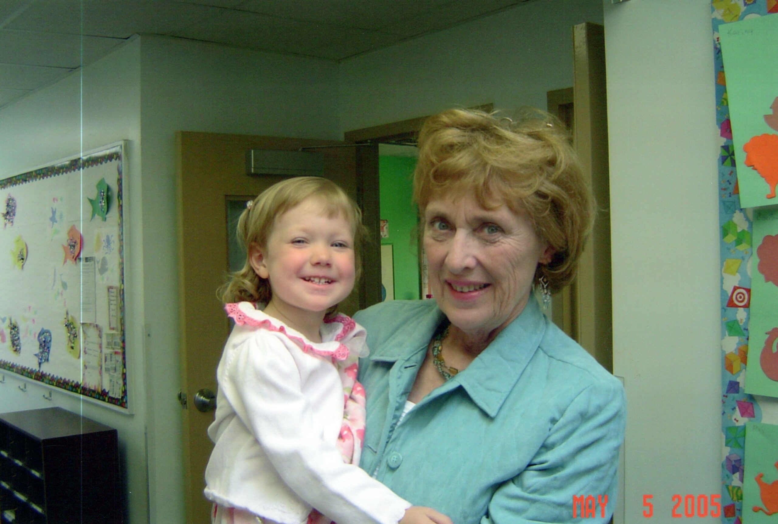 Smiling adult woman holds a young girl in a school hallway with colorful student artwork on the walls behind them.