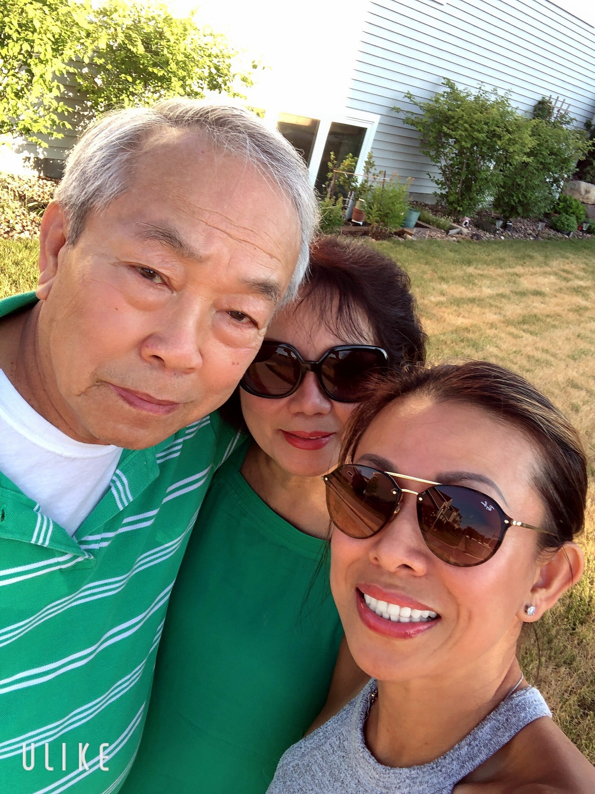 Three adults pose for a close-up outdoor selfie in a backyard garden. Man in a green striped polo, two women nearby wearing green and gray outfits with sunglasses.
