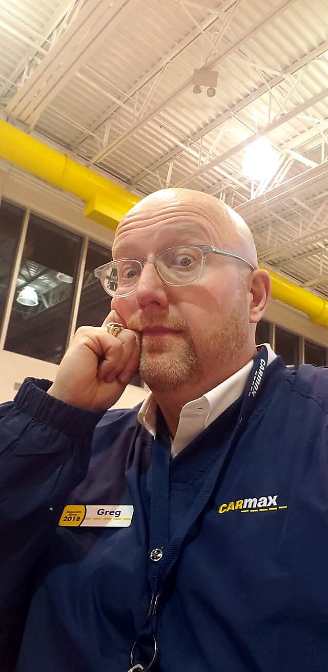 Close-up of a bald man with glasses resting his chin on his hand, wearing a CarMax uniform and name badge that reads Greg.