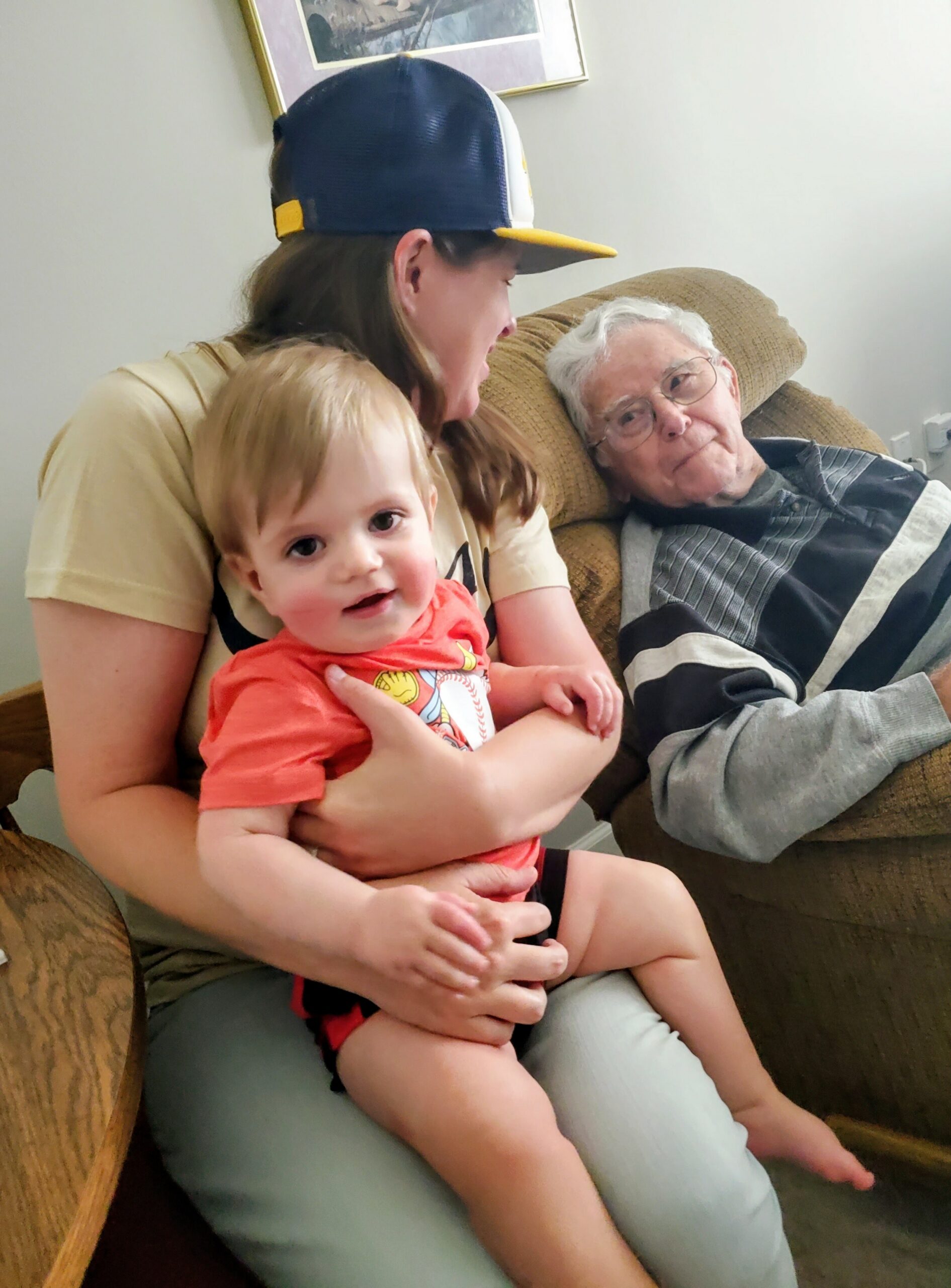 A woman in a beige shirt sits on a couch, holding a smiling toddler in a coral shirt while an elderly man in a striped sweater sits beside them on a brown recliner.