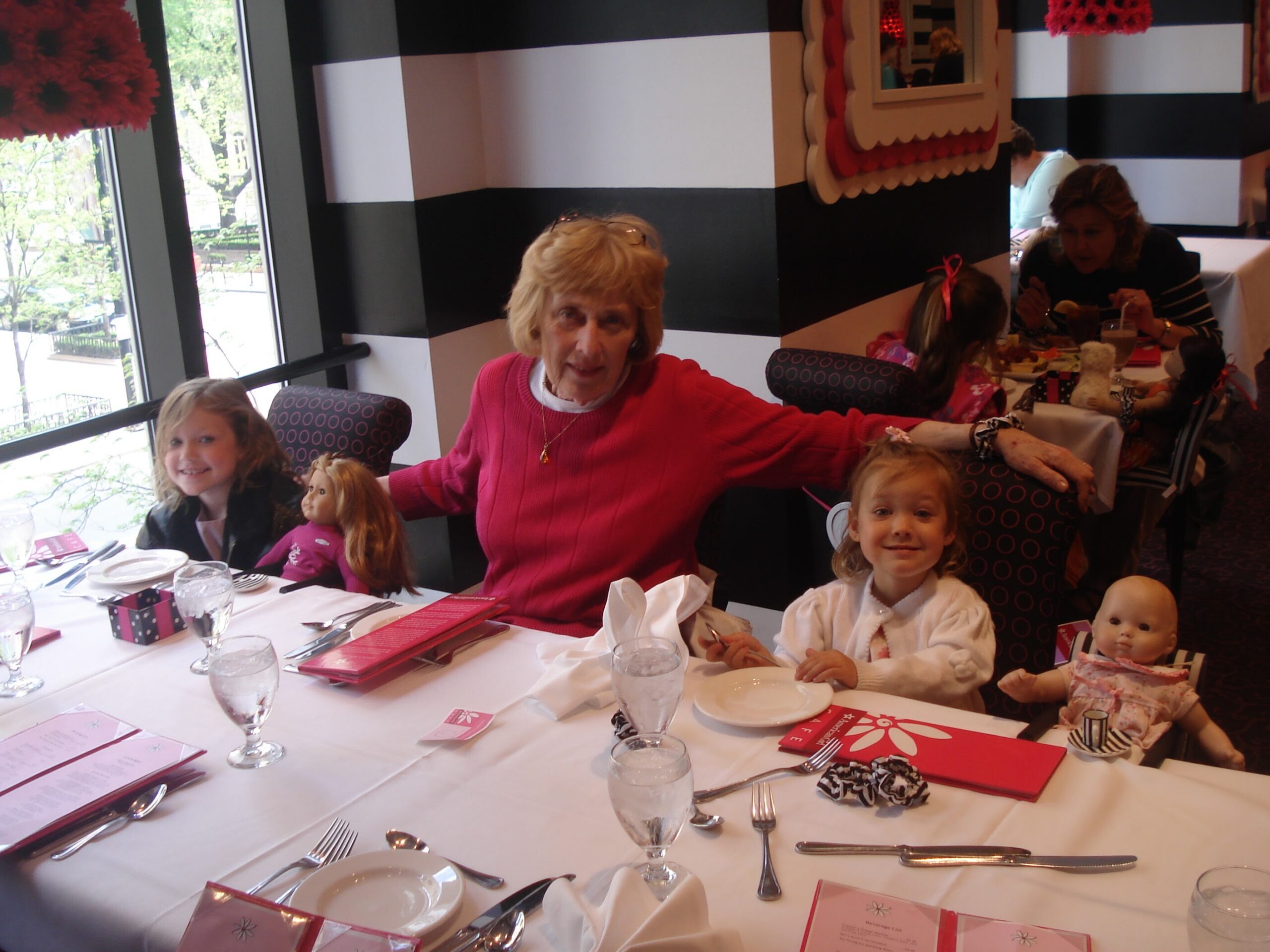 A group of people dining at a long table in a restaurant, with children and an older woman in a red sweater at center.
