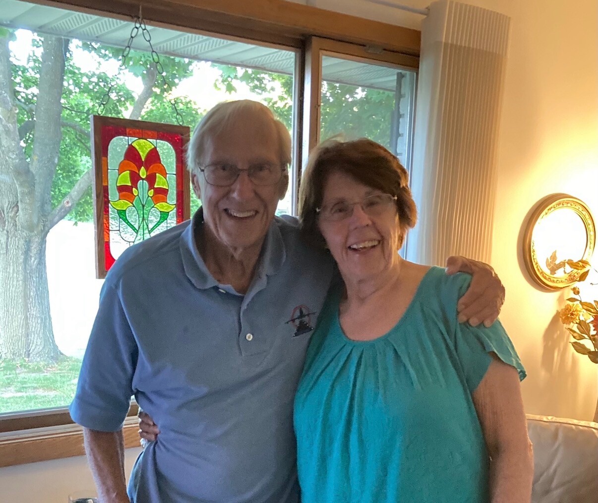 Smiling older couple stands together in a living room, arm in arm, near a window with greenery outside and a colorful stained-glass panel nearby.