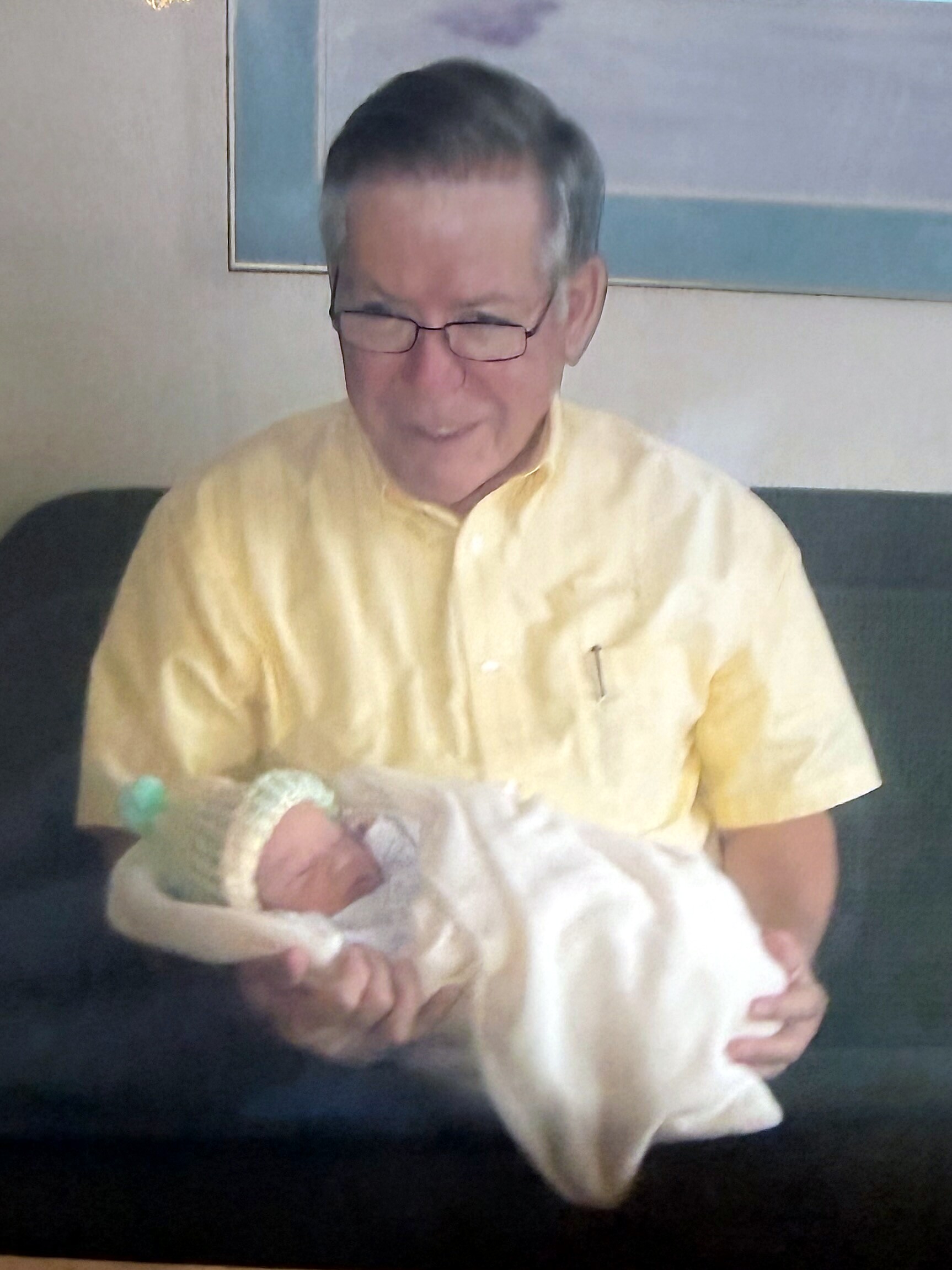 Middle-aged man with glasses in a pale yellow shirt holding a newborn baby wrapped in a white blanket on a couch.