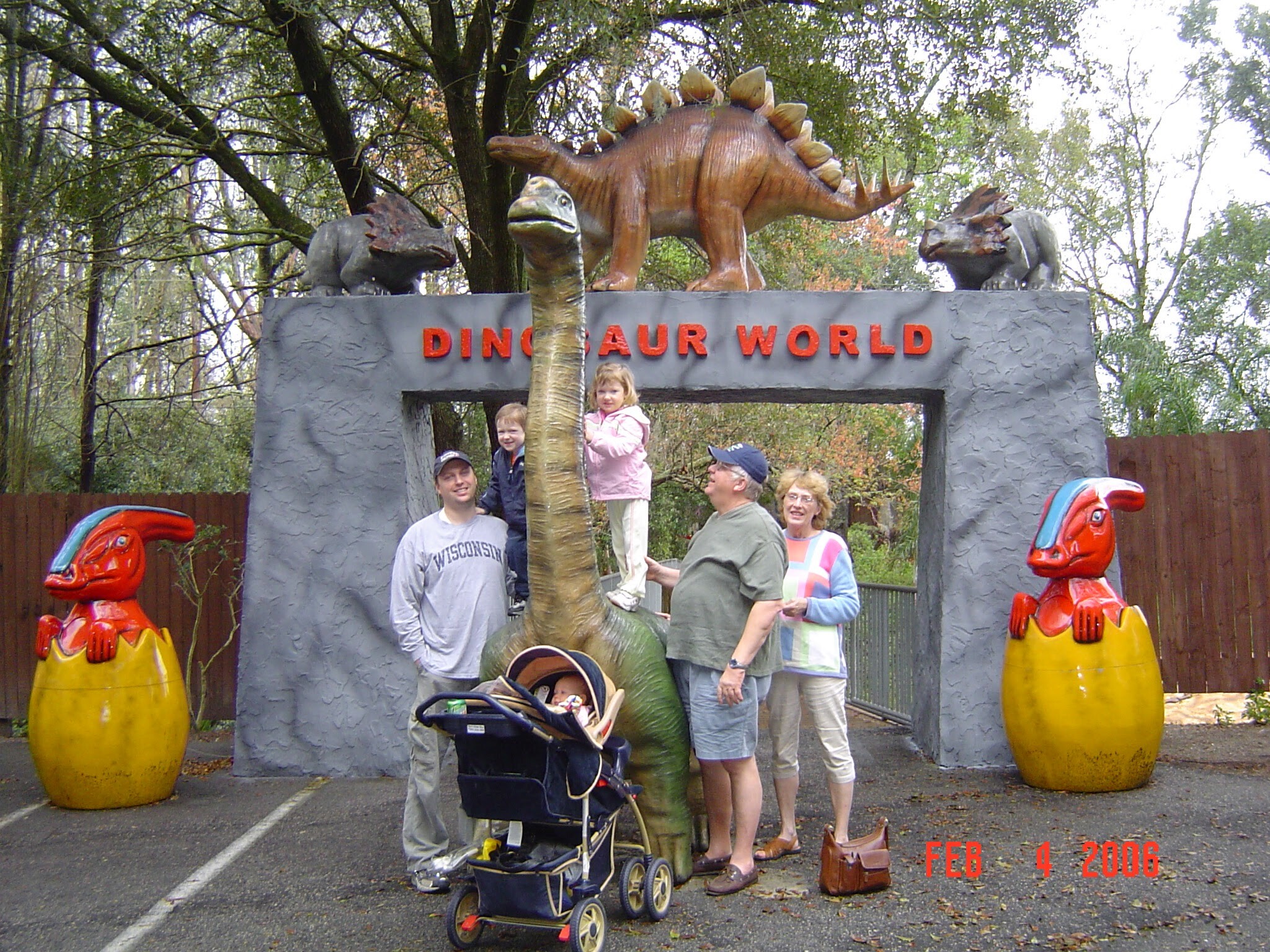 Entrance to Dinosaur World with life-size dinosaur statues and visitors posing under a gray stone arch, the sign reading 'DINOSAUR WORLD'.