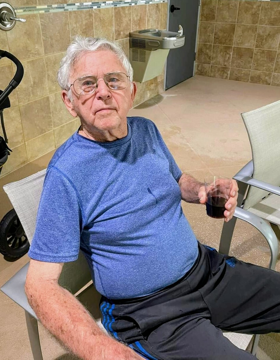 Older man with glasses wearing a blue shirt sits in a chair, holding a glass of dark beverage.
