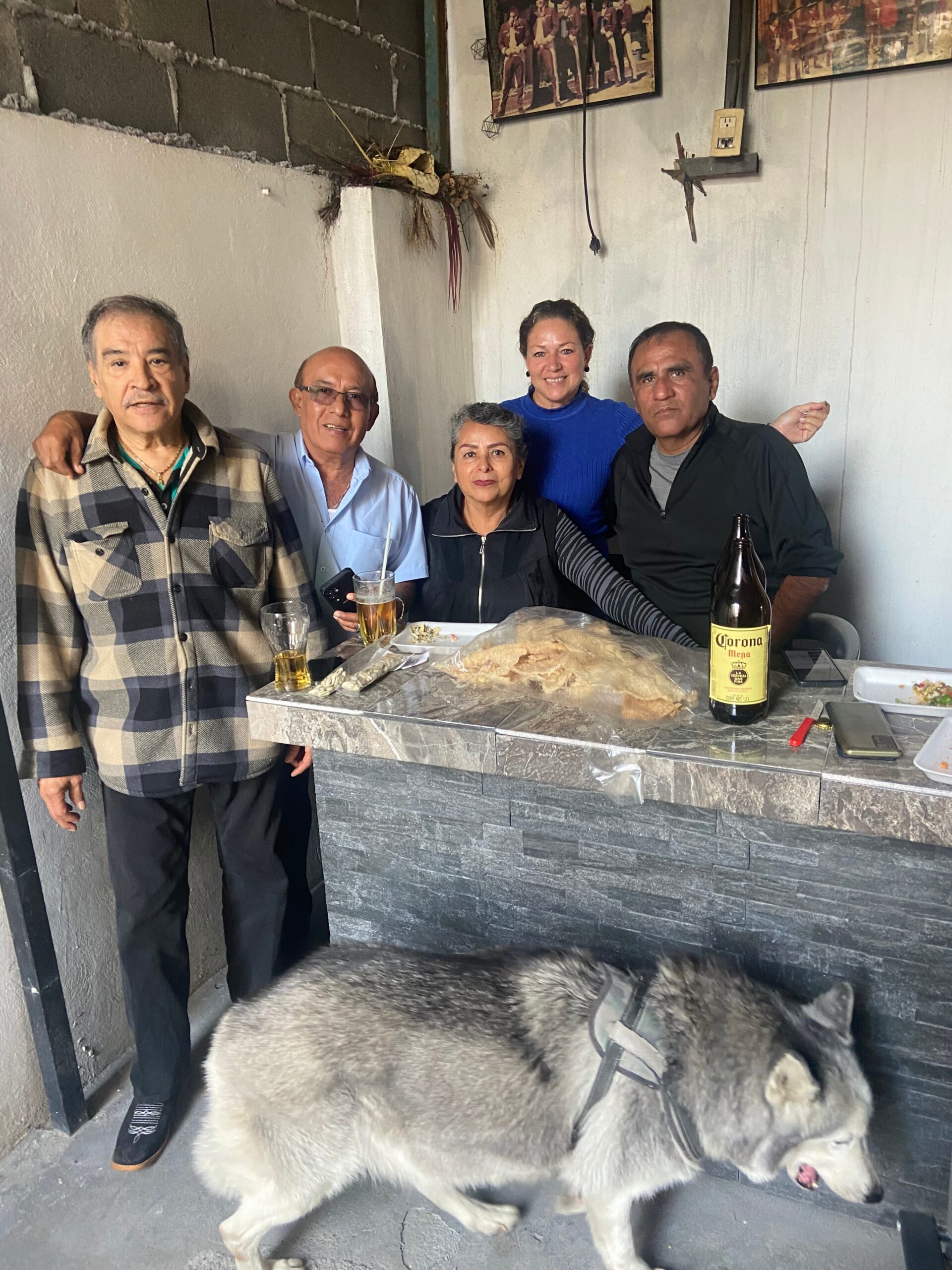 Five adults posing together indoors at a table with drinks and bread, while a large gray dog wearing a harness stands in front of them. A Corona beer bottle and plates are on the counter.