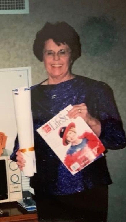 Smiling woman with short dark hair and glasses in a blue sparkly top, holding a rolled certificate and a magazine with a red-hatted figure on the cover.