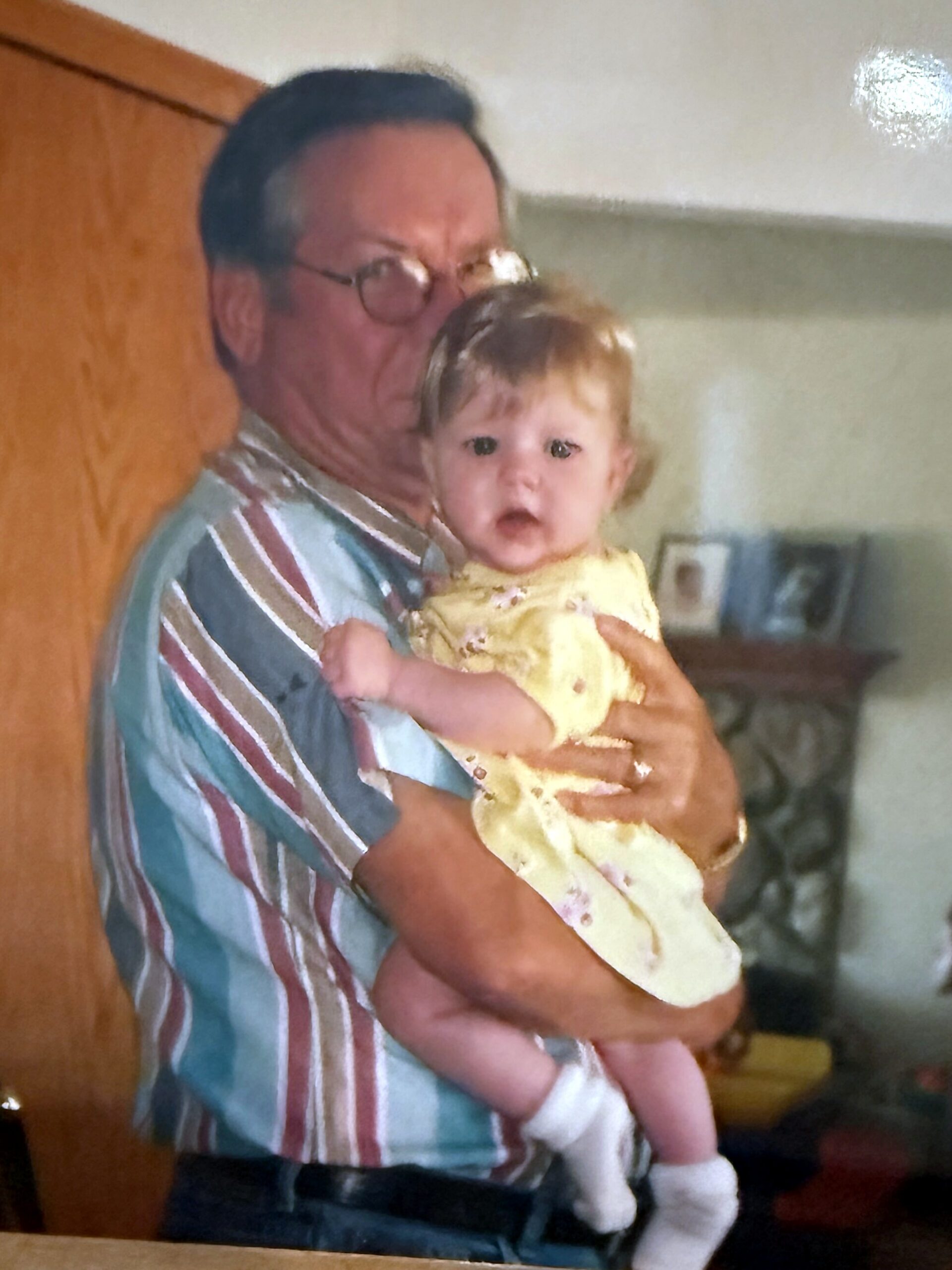 Older man with glasses holding a baby girl indoors, both facing the camera.