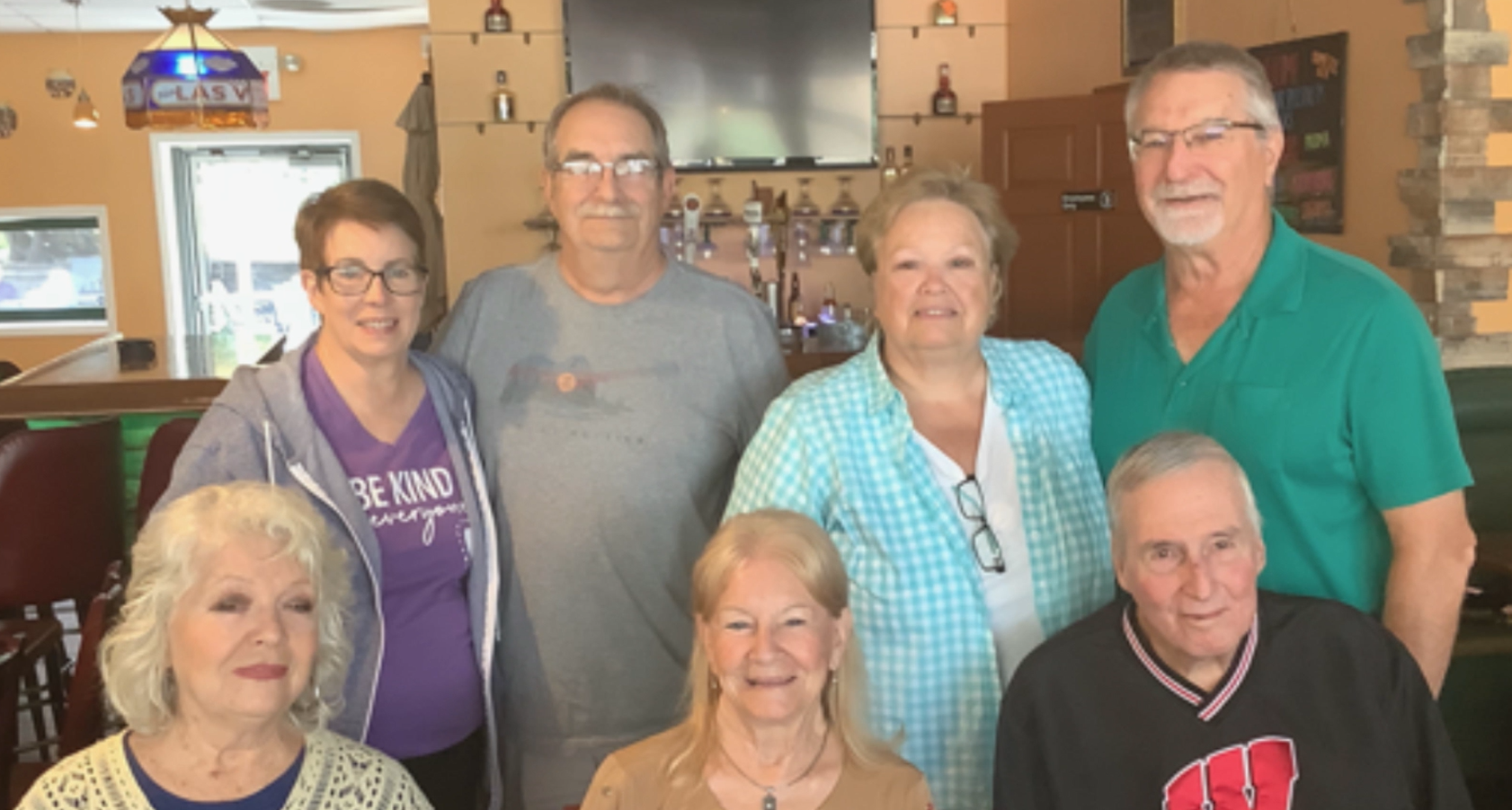 Seven seniors posing together in a cozy bar/restaurant interior with bottles on display behind them.