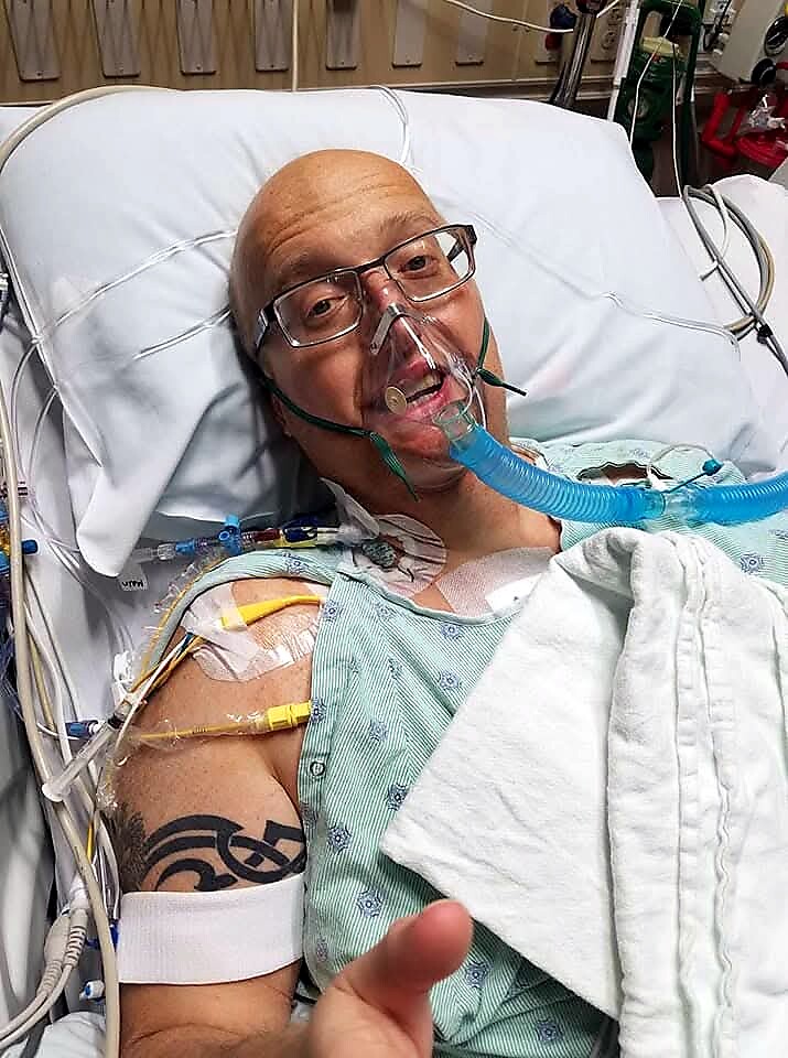 HospitaI patient lying in bed wearing an oxygen mask, connected to tubes and monitors, with a visible tattoo on the left arm.