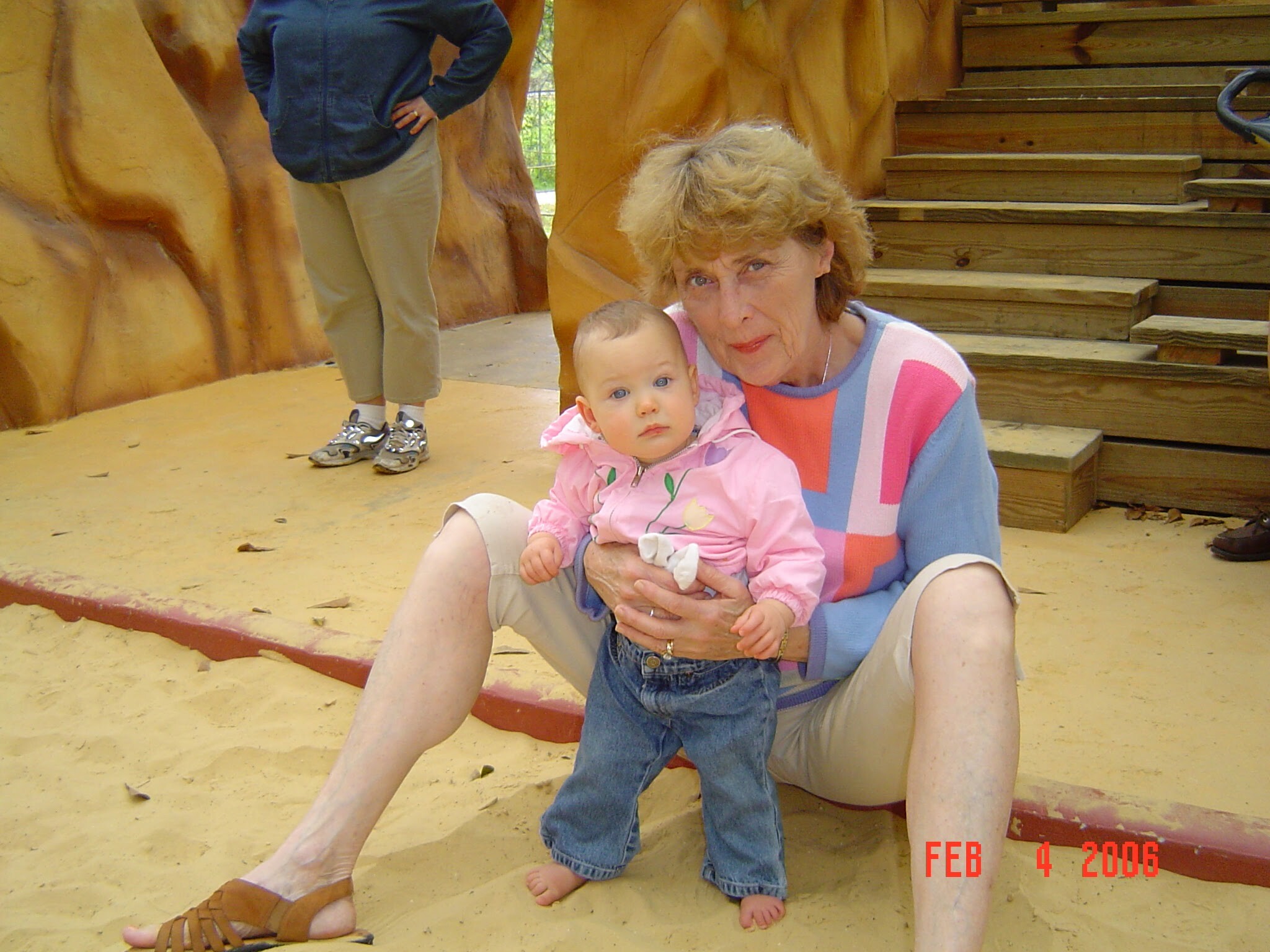 Grandmother with short light hair holding a baby girl in a pink jacket on a sandy outdoor area, with wooden steps behind them.
