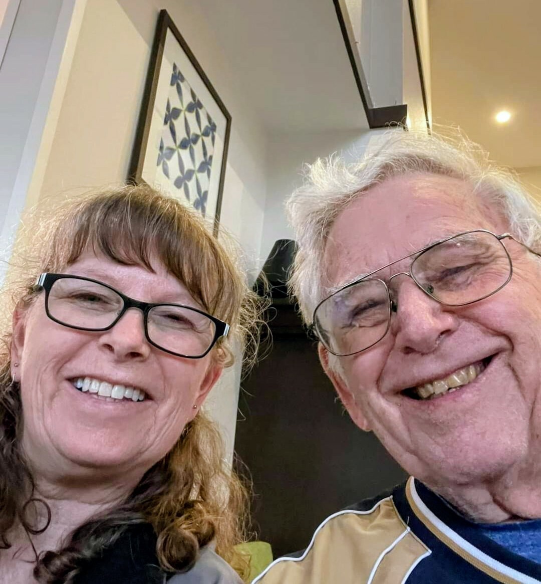 Two smiling adults pose for a selfie indoors, looking at the camera. The woman on the left wears glasses and has light brown hair; the man on the right has white hair and wears glasses, both showing big smiles.