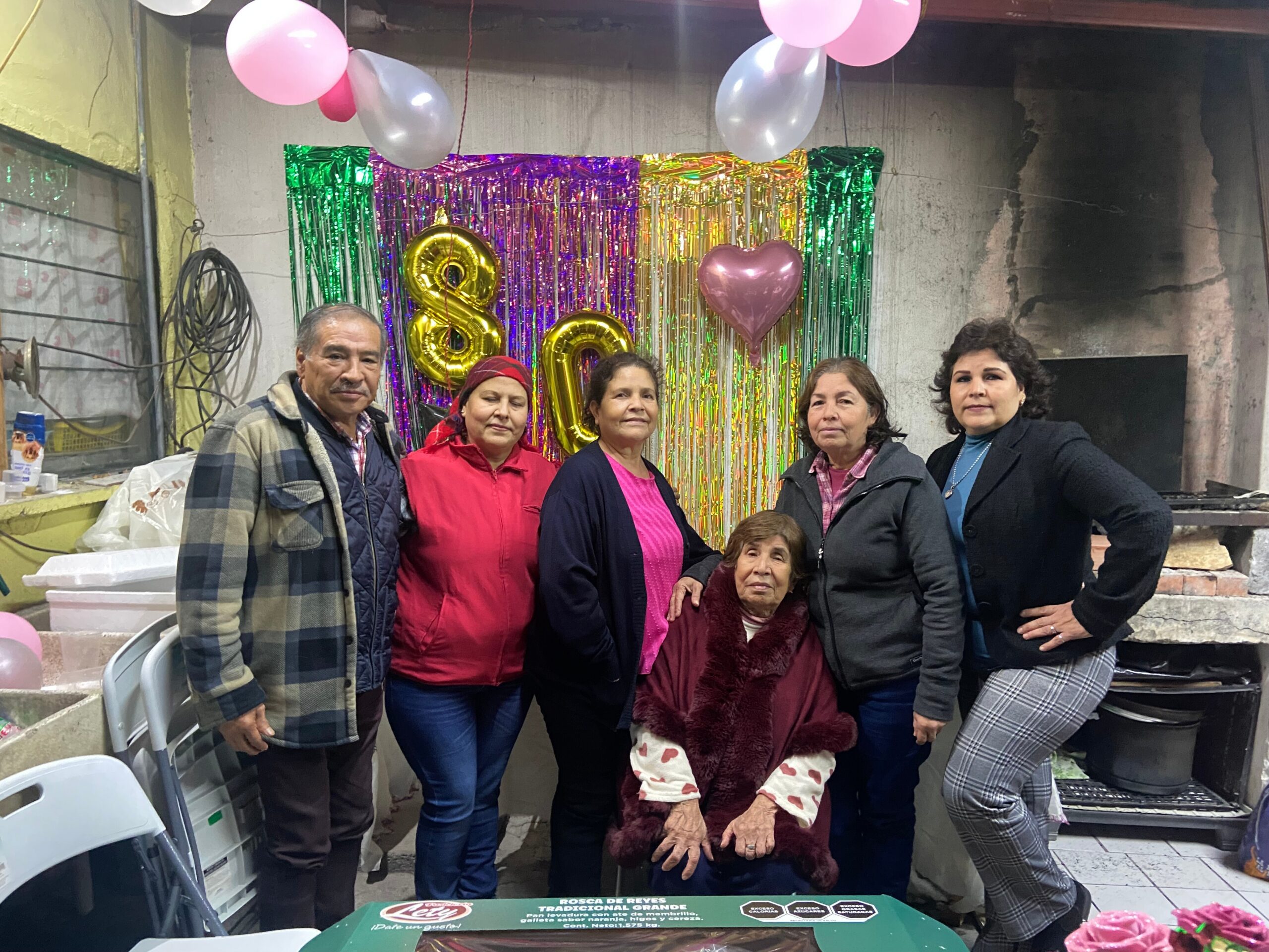 Group of six adults and one child posing for a celebration photo against a pink/foil party backdrop with balloons and heart-shaped balloon accents.