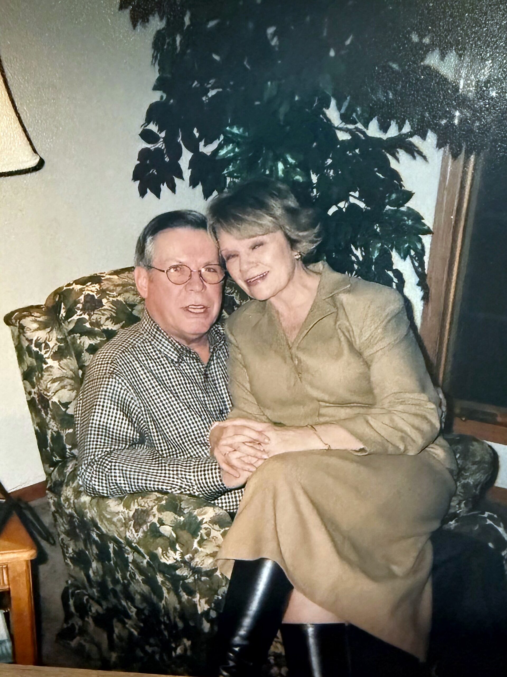 Older couple sitting close together on a floral sofa, smiling at the camera in a living room setting.