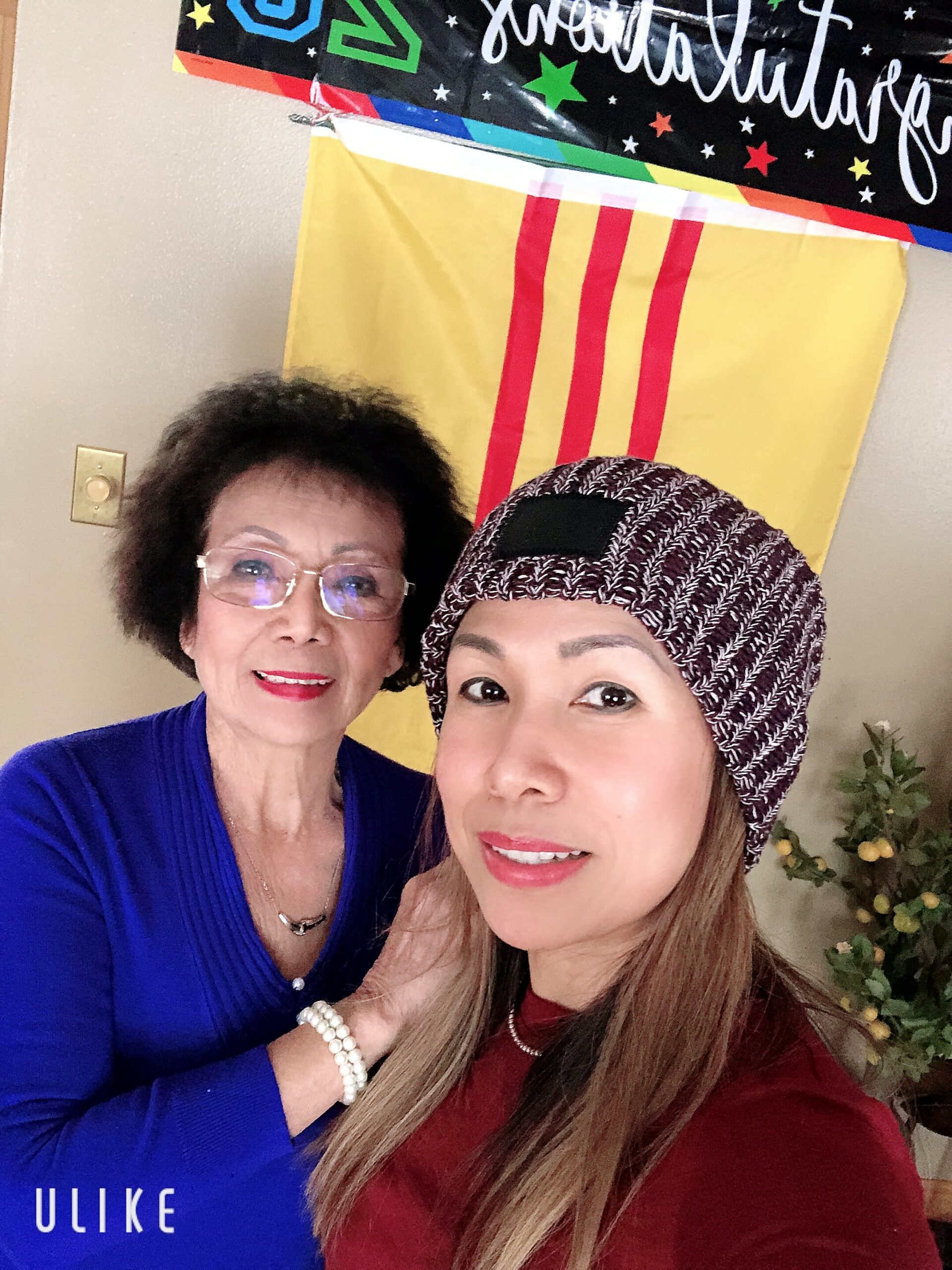 Two women pose for a selfie indoors, one older in a blue top with pearl bracelets and glasses, the other younger wearing a patterned headwrap and maroon top, both smiling in front of colorful banners on the wall.