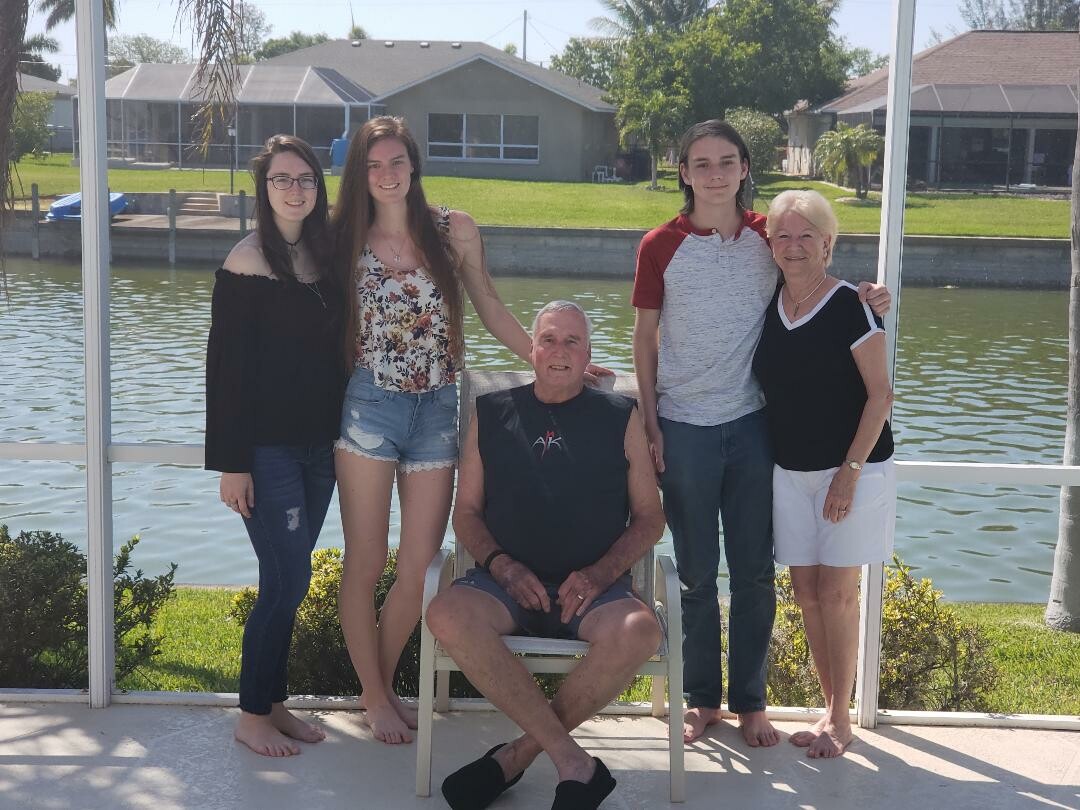 Five family members pose outdoors by a canal, with houses in the background; a man sits in a chair center among the group.