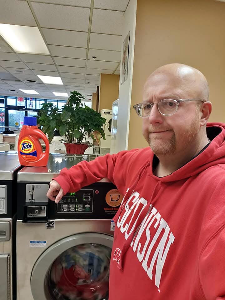 Man with glasses and a red hoodie stands at a front-loading washer in a laundromat, with Tide detergent on top of the machine, smiling at the camera.