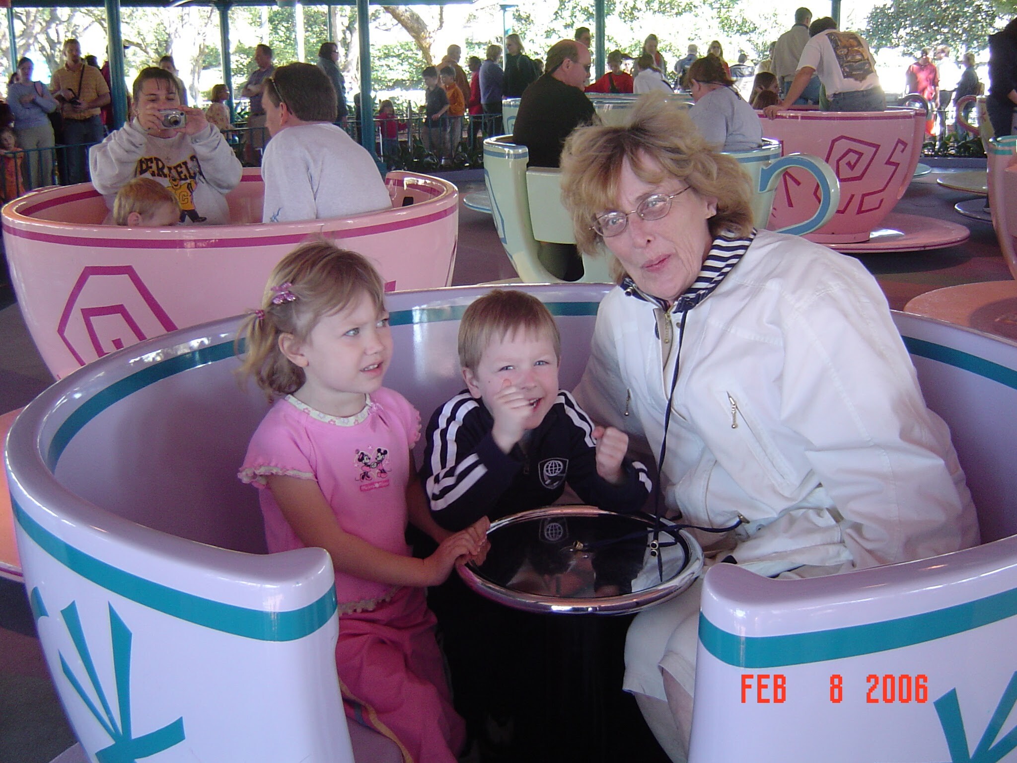 Group of children and an adult woman ride pink teacups at a crowded amusement park ride, smiling and taking photos in the background.
