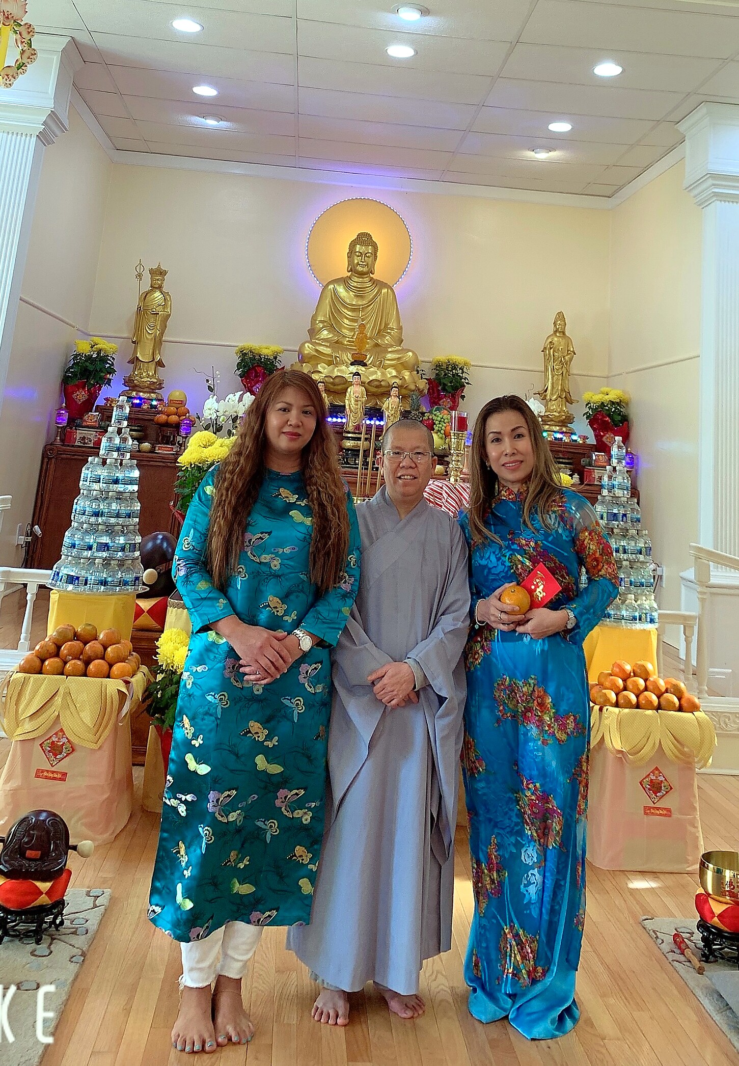 Three people in colorful áo dài pose in front of a Buddhist altar with a large golden Buddha statue, surrounded by flowers, smaller statues, and fruit offerings.