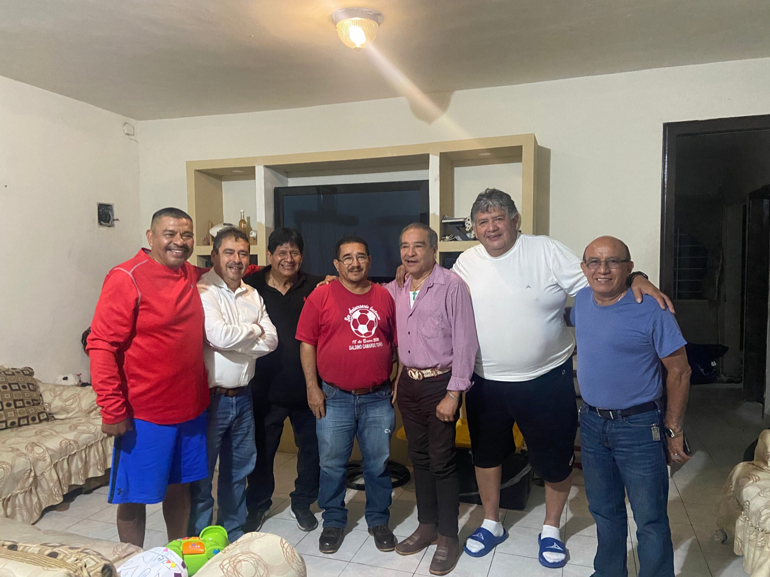 Group of men standing together indoors, smiling for the camera in a casual living-room setting.