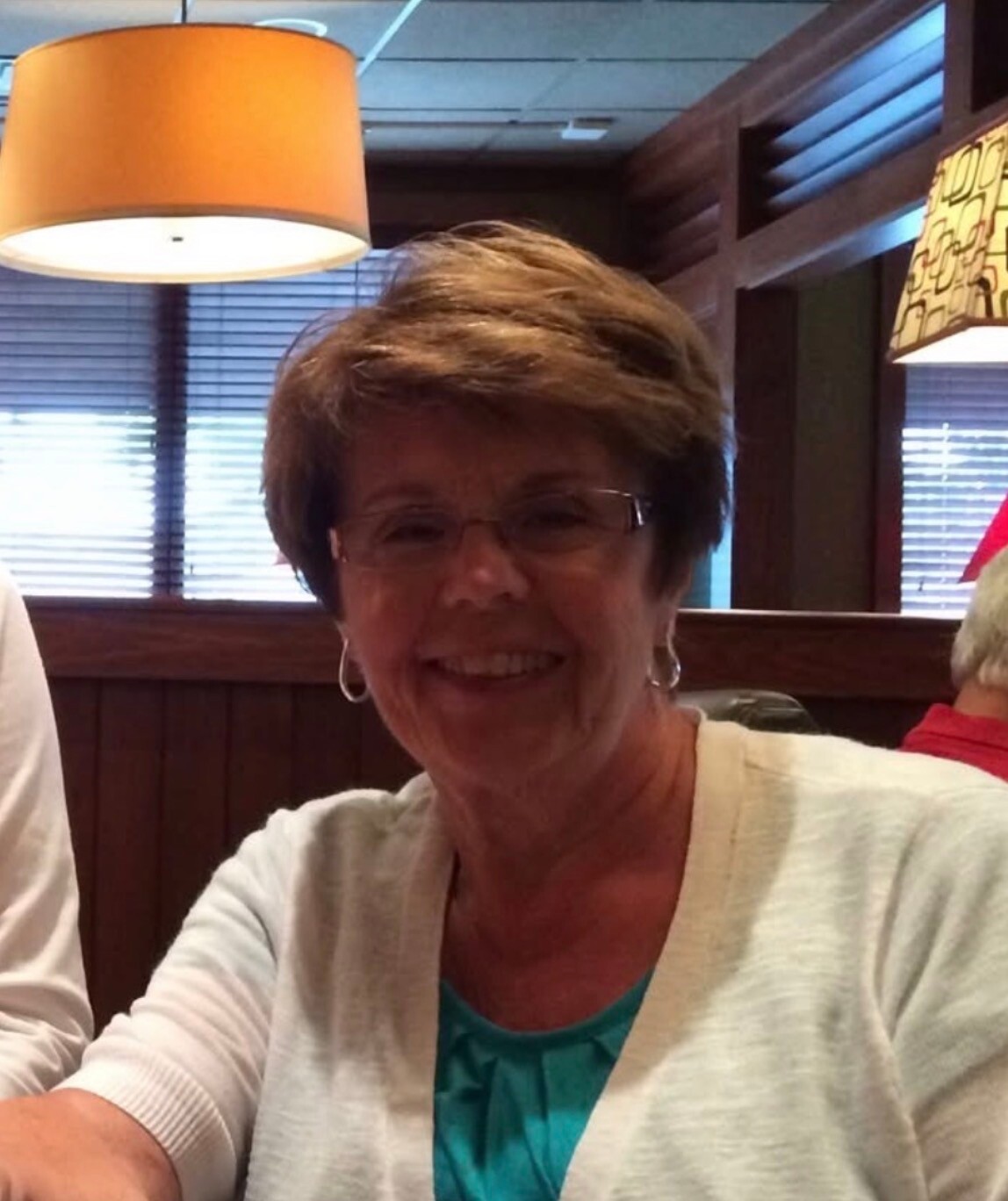 Smiling older woman with short light brown hair and glasses wearing a white cardigan over a turquoise top, seated in a restaurant with warm lighting.