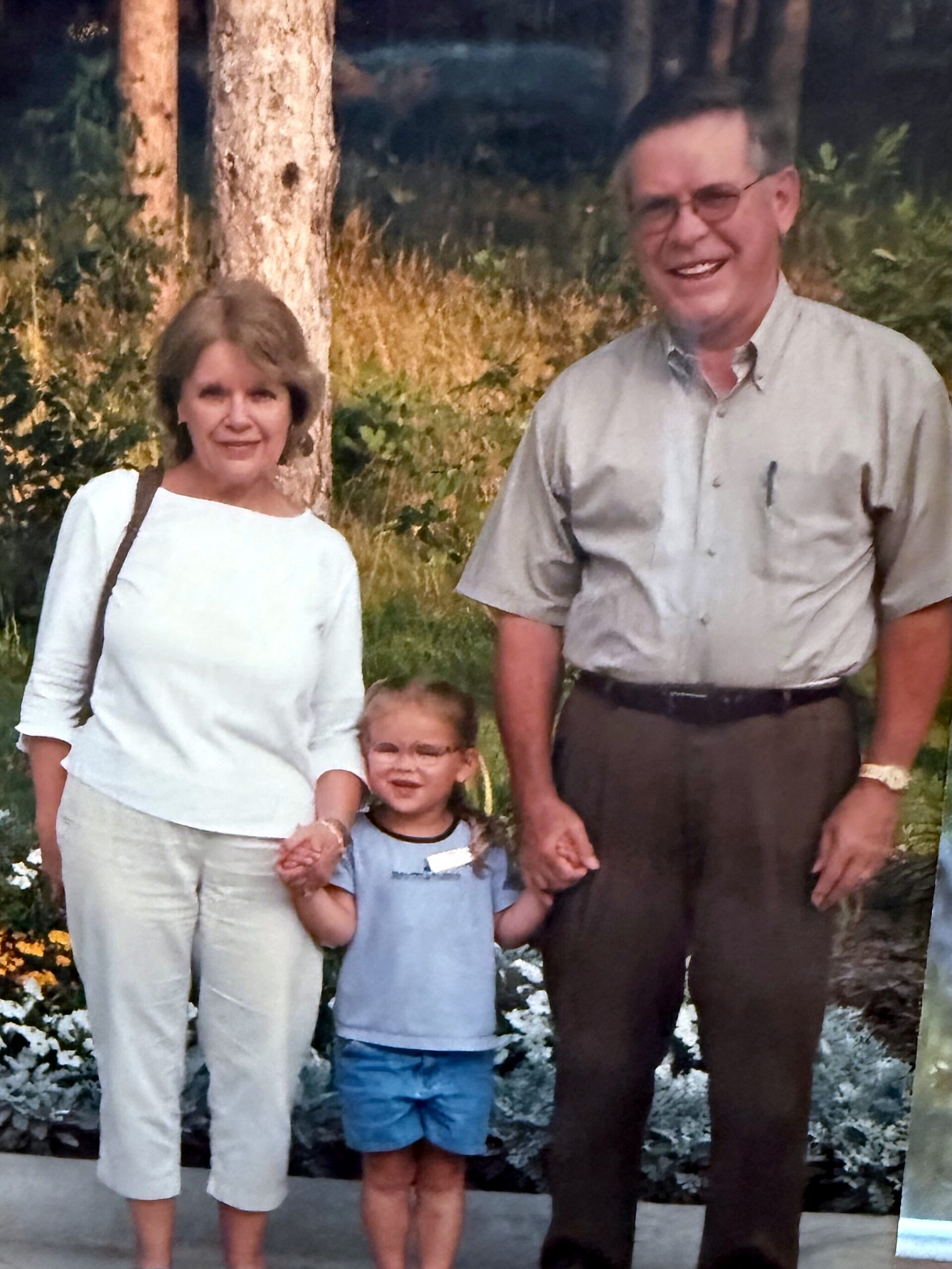 Family portrait outdoors: mother, father, and young child holding hands and smiling near trees and greenery in a sunny setting.