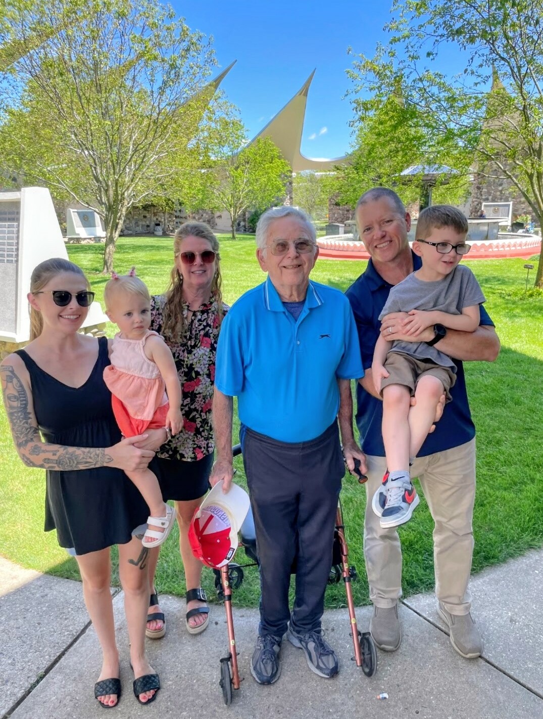 Multi-generational group of eight posing for a photo in a sunny park, elderly man with a walker at center, smiling family members around him.