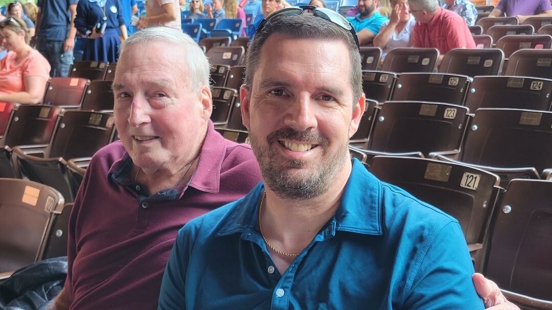Two men smiling at the camera while seated in a stadium with brown chairs and other attendees in the background.