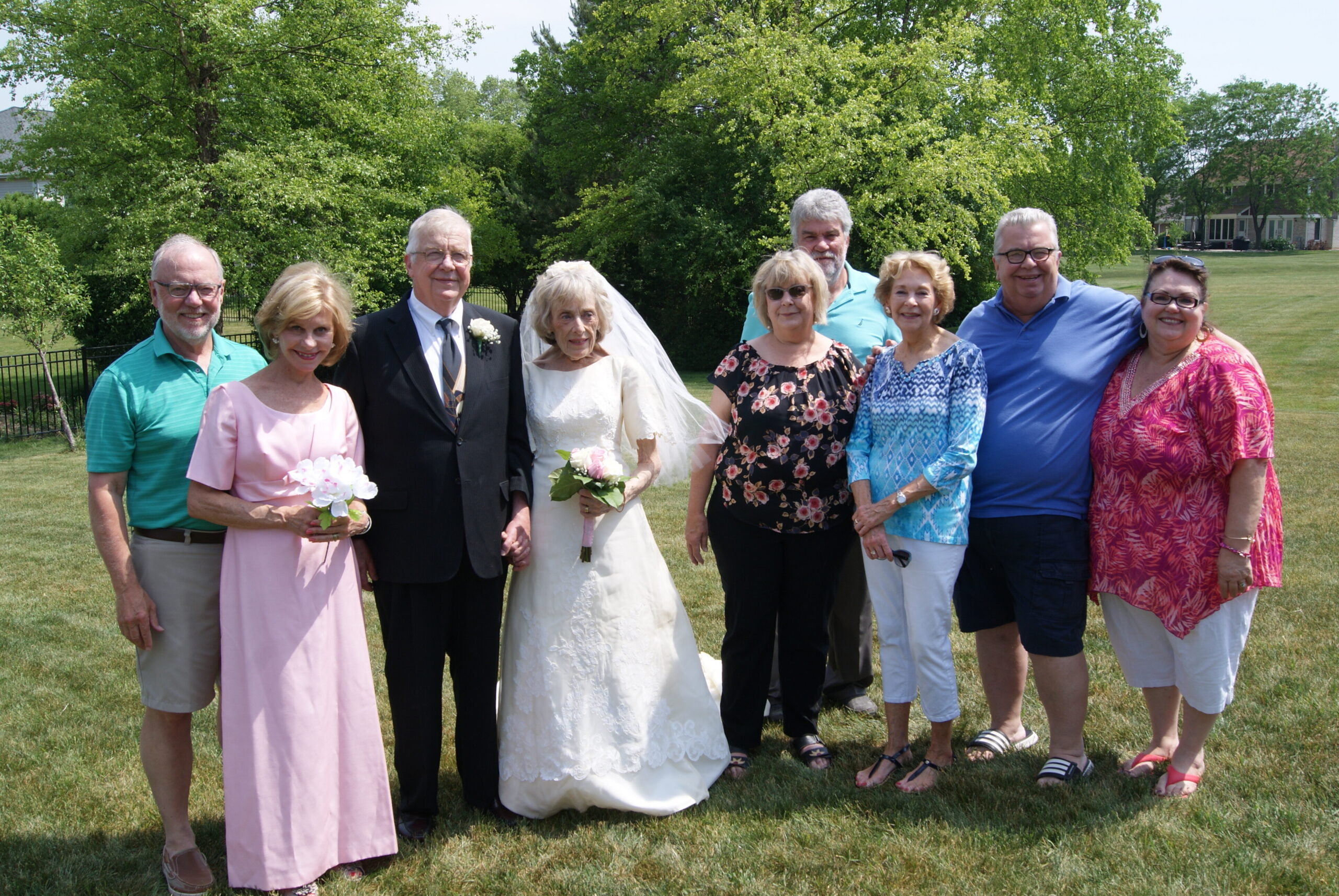 Group of wedding guests posing together on a sunny lawn, with the bride and groom in the center and smiling friends surrounding them.
