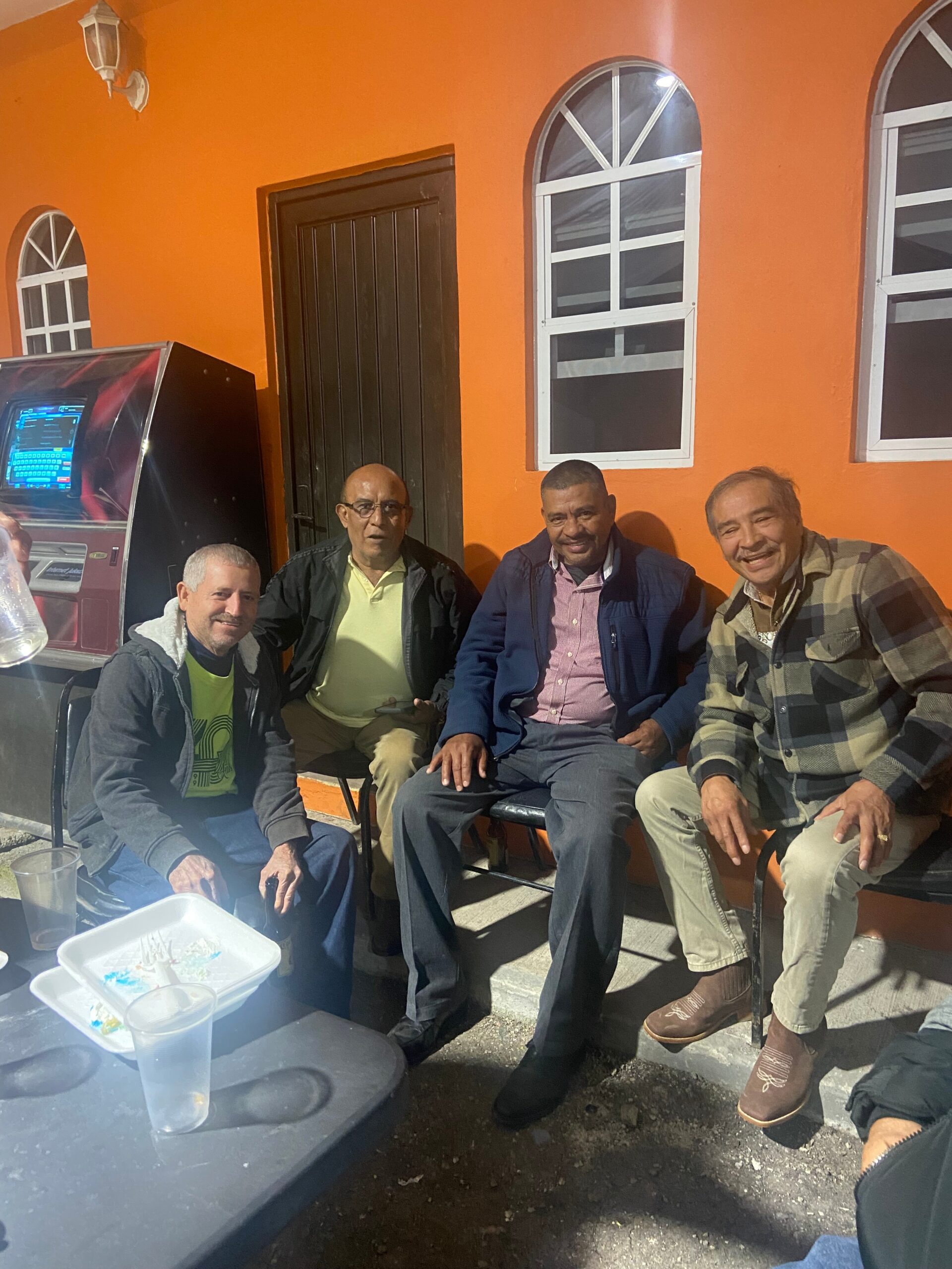 Four men sit together outside an orange building with arched white-trimmed windows, smiling at the camera.
