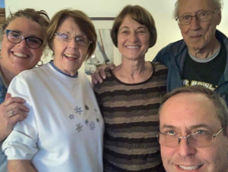 Five adults smiling for a group selfie indoors, with a man in the foreground taking the photo and others gathered closely behind him.