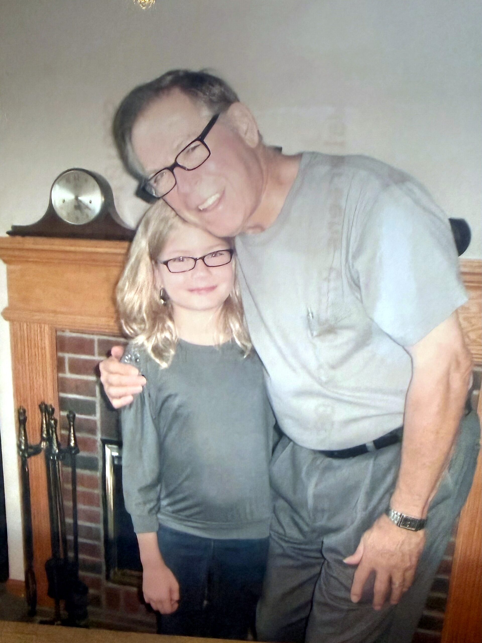 Older man and young girl pose together with heads touching in a living room near a brick fireplace, both wearing glasses and gray tops.