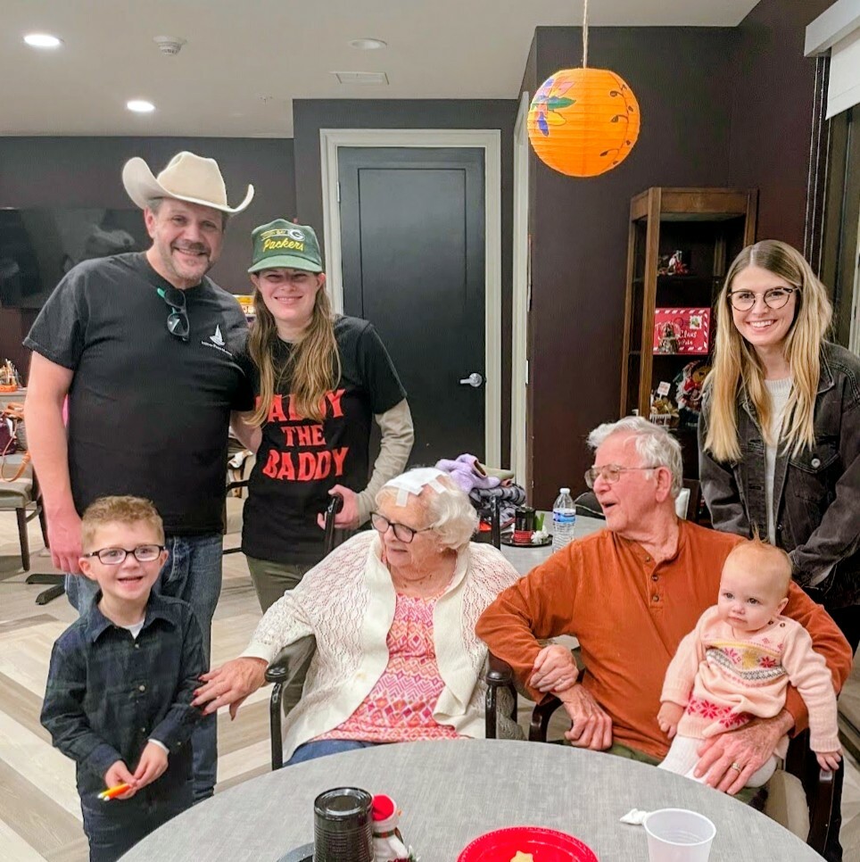 A multigenerational group posing for a photo at a party: an elderly woman in a wheelchair, a man with an orange shirt holding a baby, a boy with glasses, and three younger adults smiling nearby in a cozy indoor room with an orange lantern overhead.