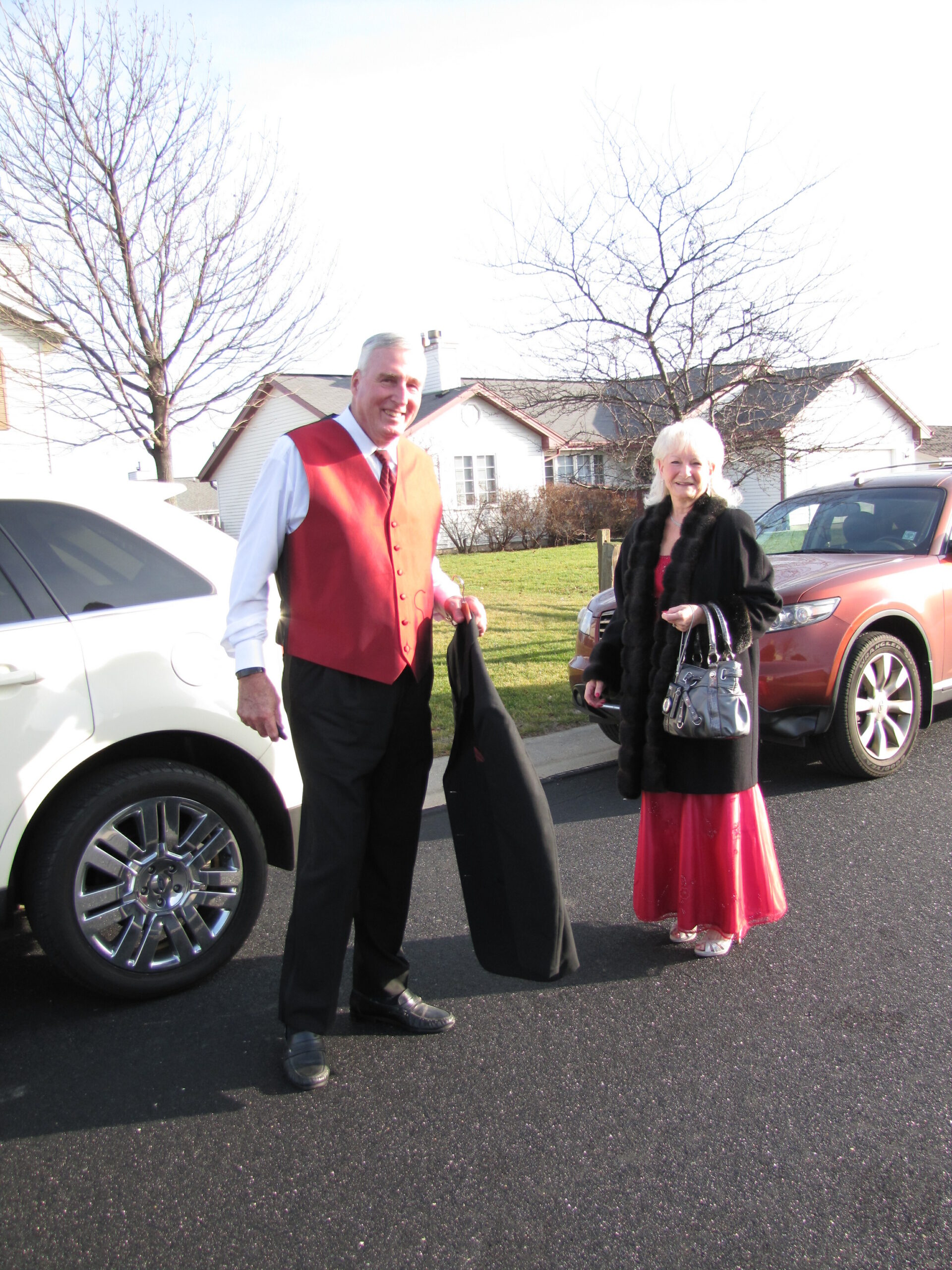 Smiling man in a red vest and white shirt stands beside a white SUV, holding a black coat; a woman in a red dress with a black fur coat stands nearby in a driveway.