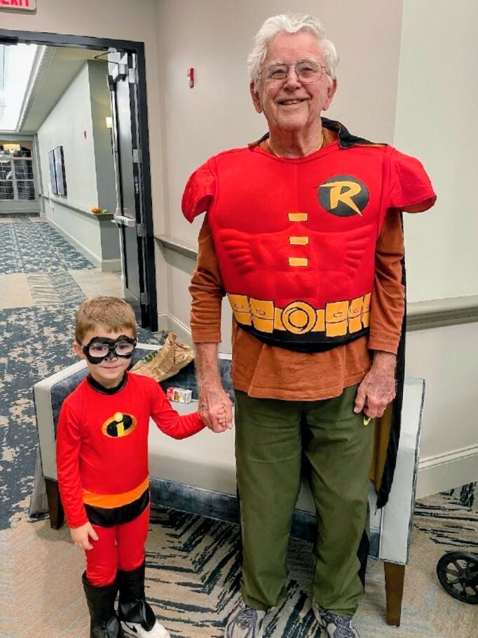 Older man and a young boy dressed as Mr. Incredible pose and hold hands in a hallway.