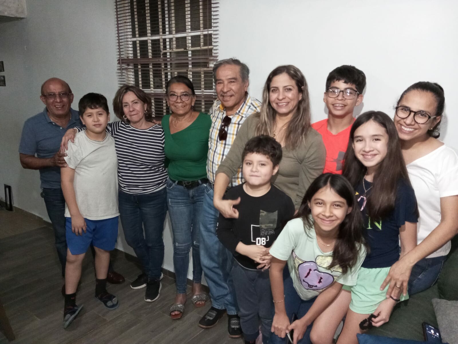Group of ten people of varying ages standing together indoors, smiling at the camera. Behind them is a window with blinds and a plain wall.