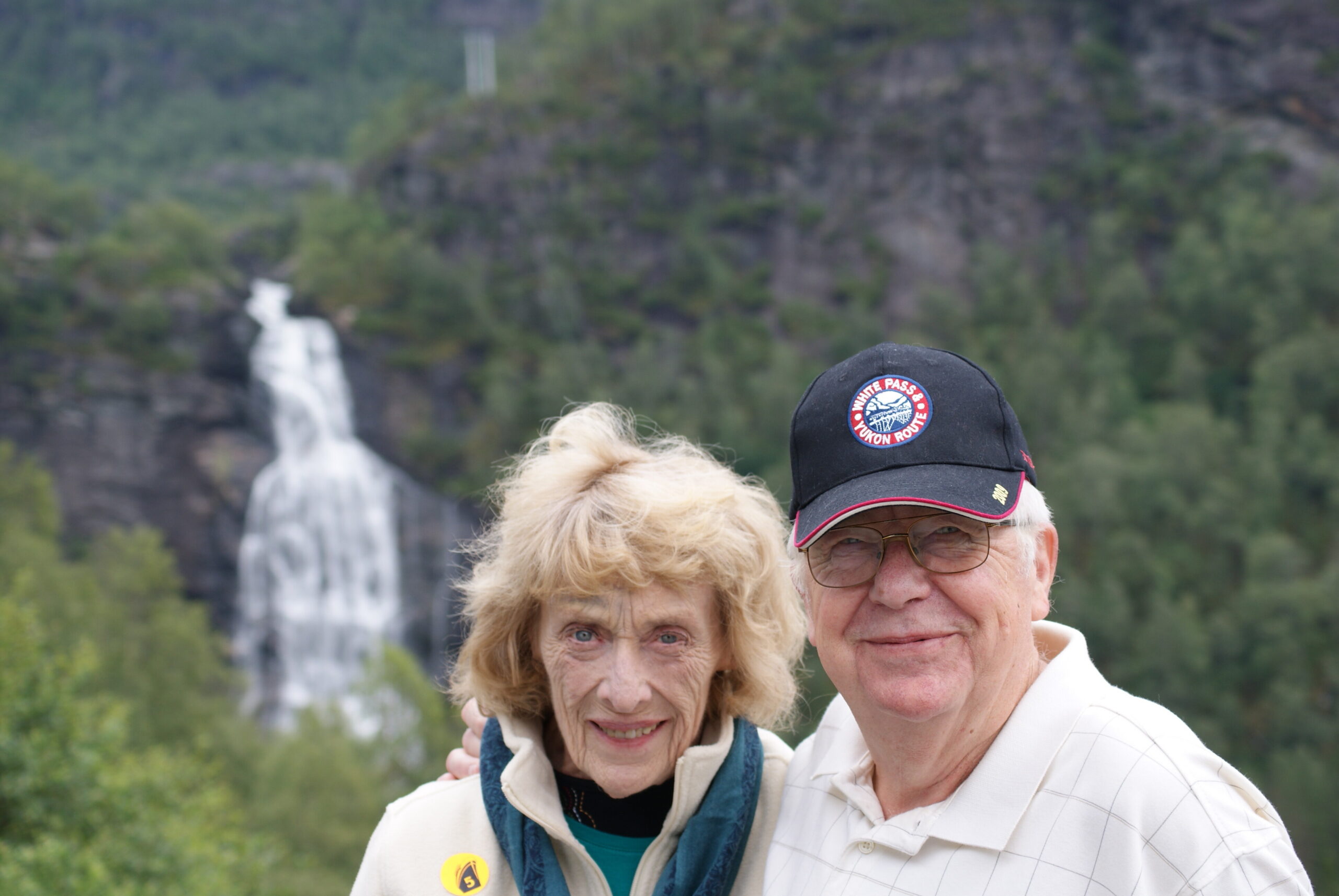Two smiling seniors posing outdoors with a waterfall and mountains behind them, suggesting a scenic tourist spot.