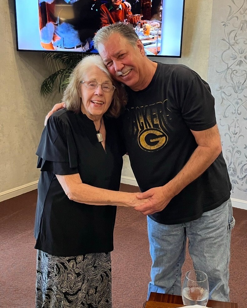 Smiling elderly woman and a man hug in a living room, TV on wall behind them.